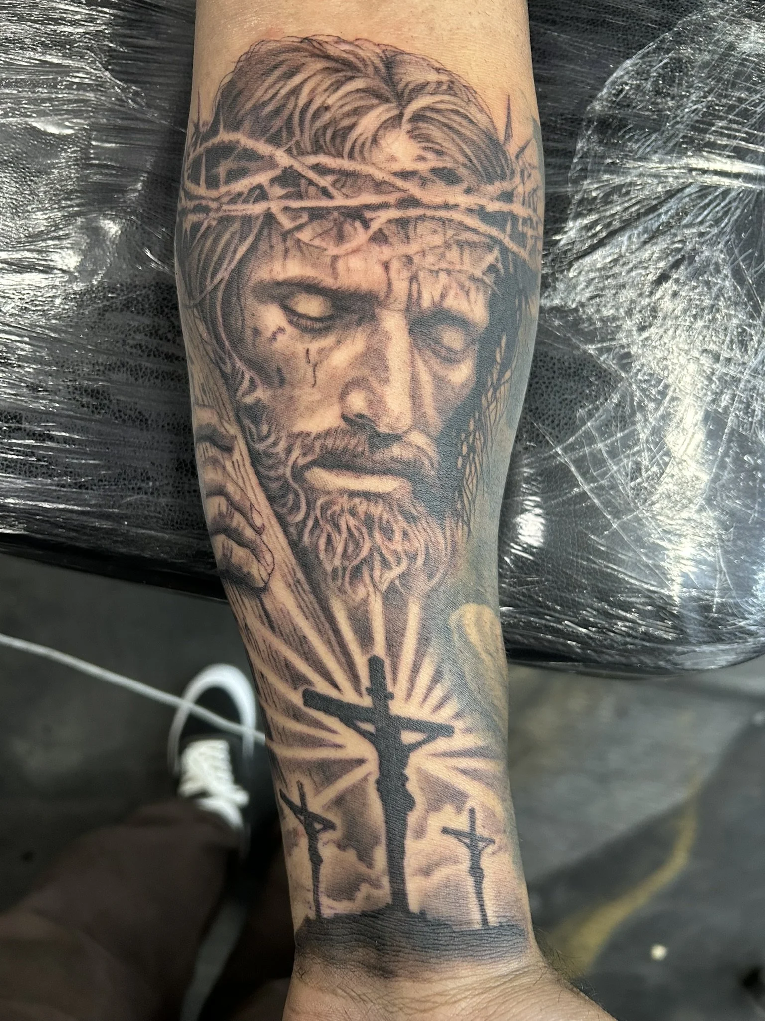 Detailed black-and-gray forearm tattoo featuring a distressed bearded man wearing a crown of thorns, with a cross and radiating light below.