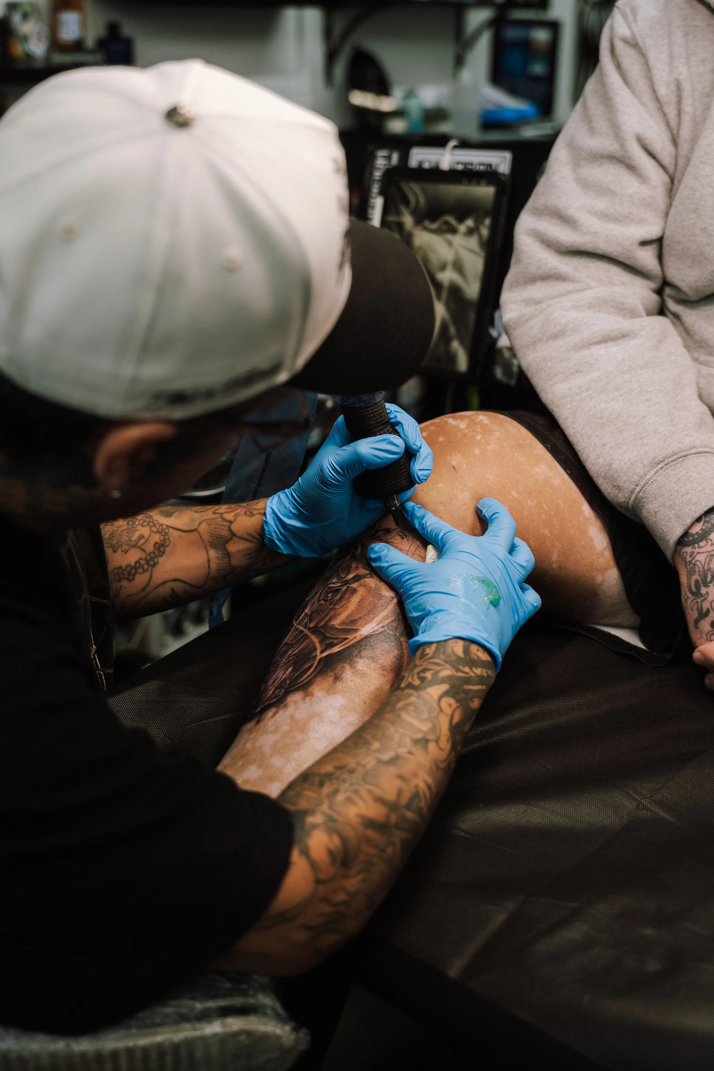 Tattoo artist wearing blue gloves works on a client’s tattooed arm with a tattoo machine in a studio.