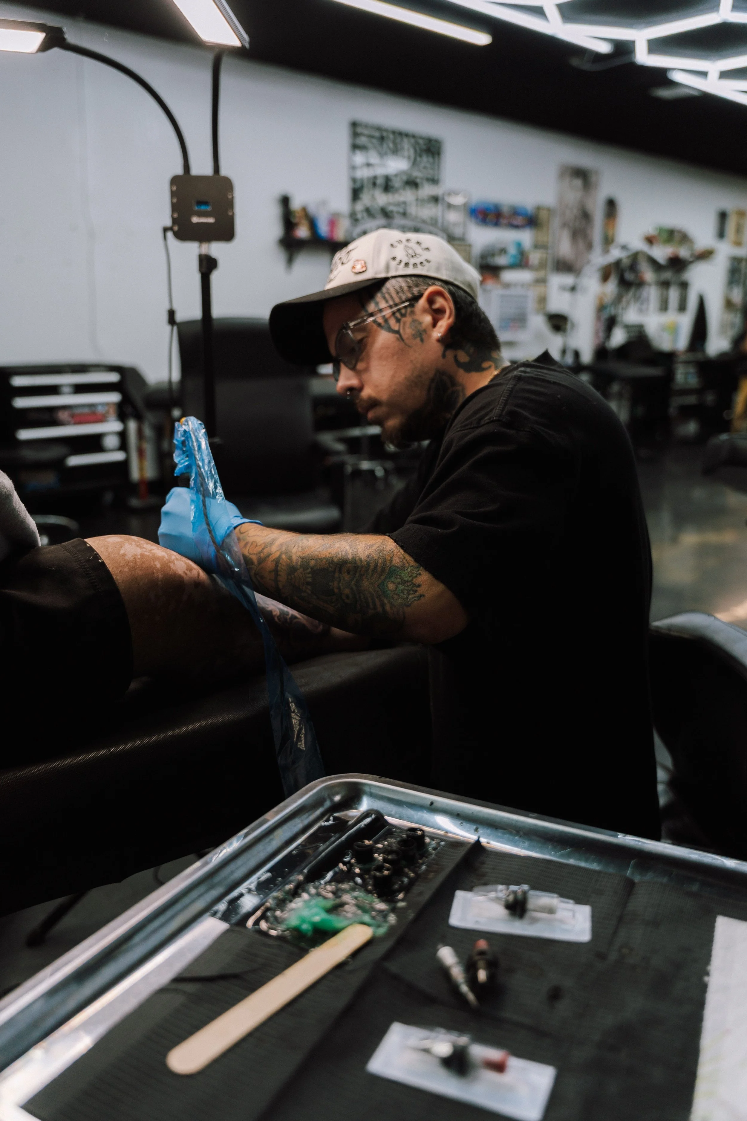 Tattooed artist wearing a cap and glasses works in a tattoo studio beside a tray of tattoo equipment.