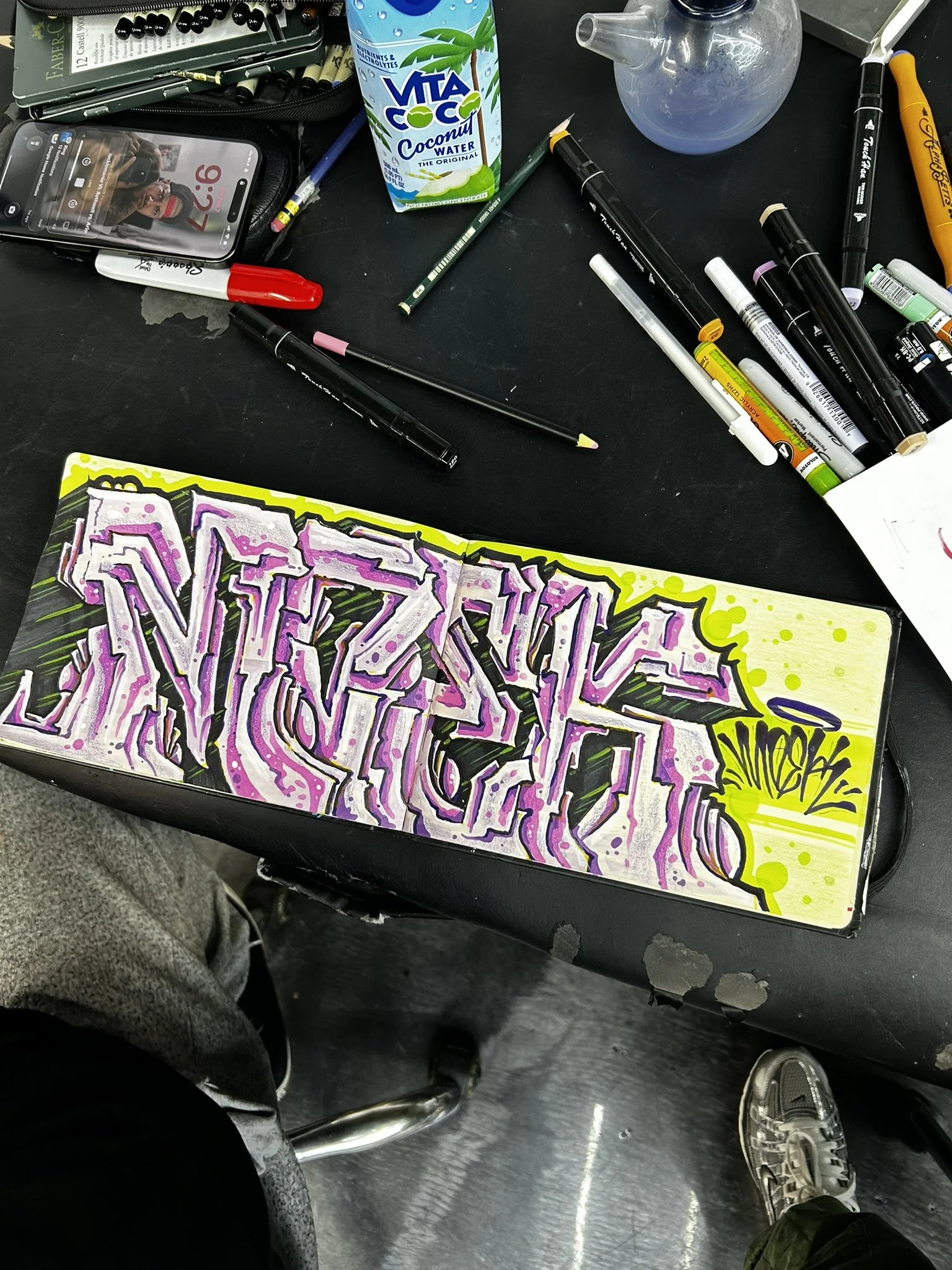 Graffiti artwork in progress surrounded by paint markers, pencils, a smartphone, coconut water, and art supplies on a black worktable.