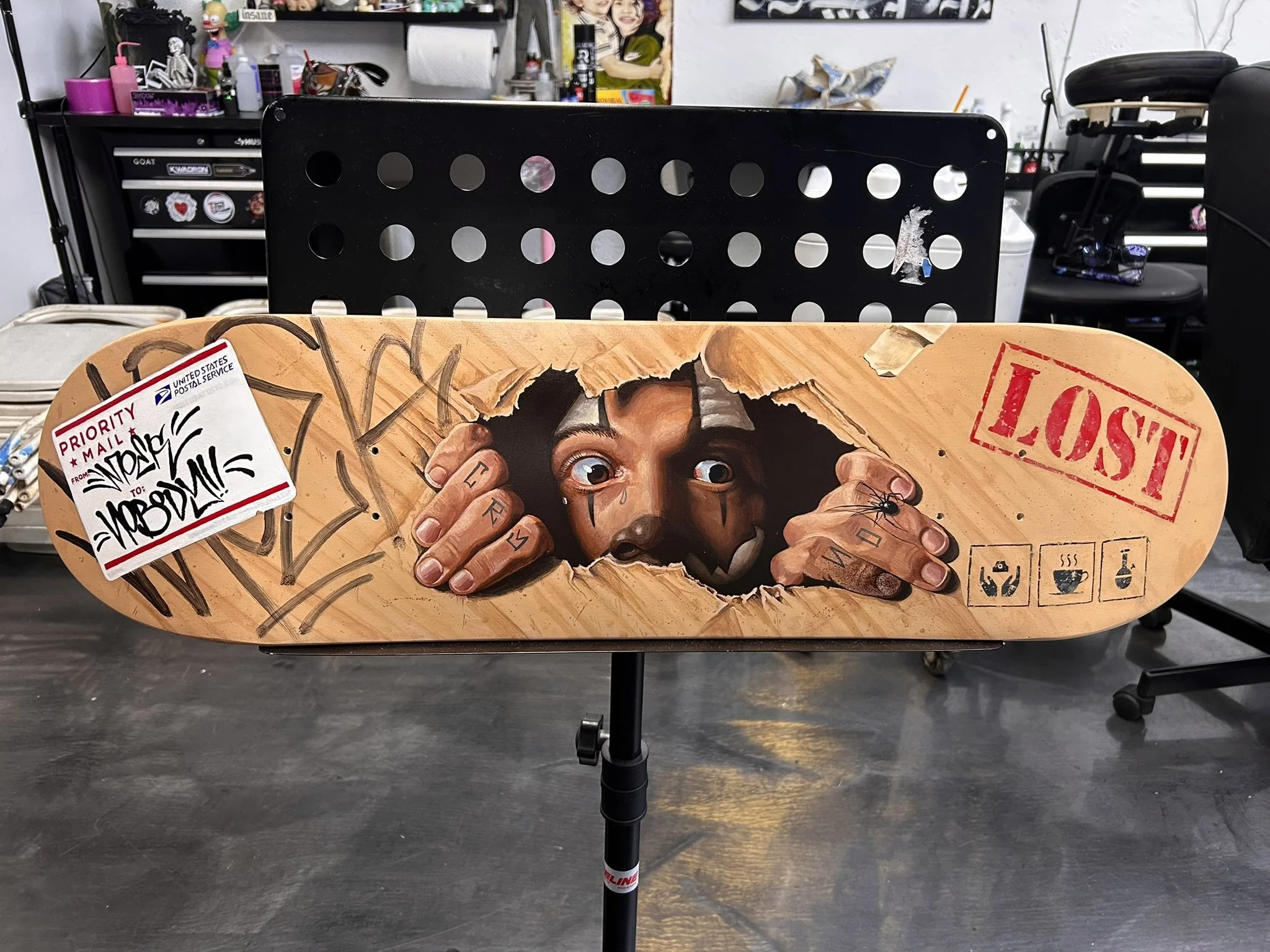 Decorated skateboard deck featuring a crying man breaking through wood, graffiti, postal mail artwork, a spider, and a red “LOST” stamp in an art studio.