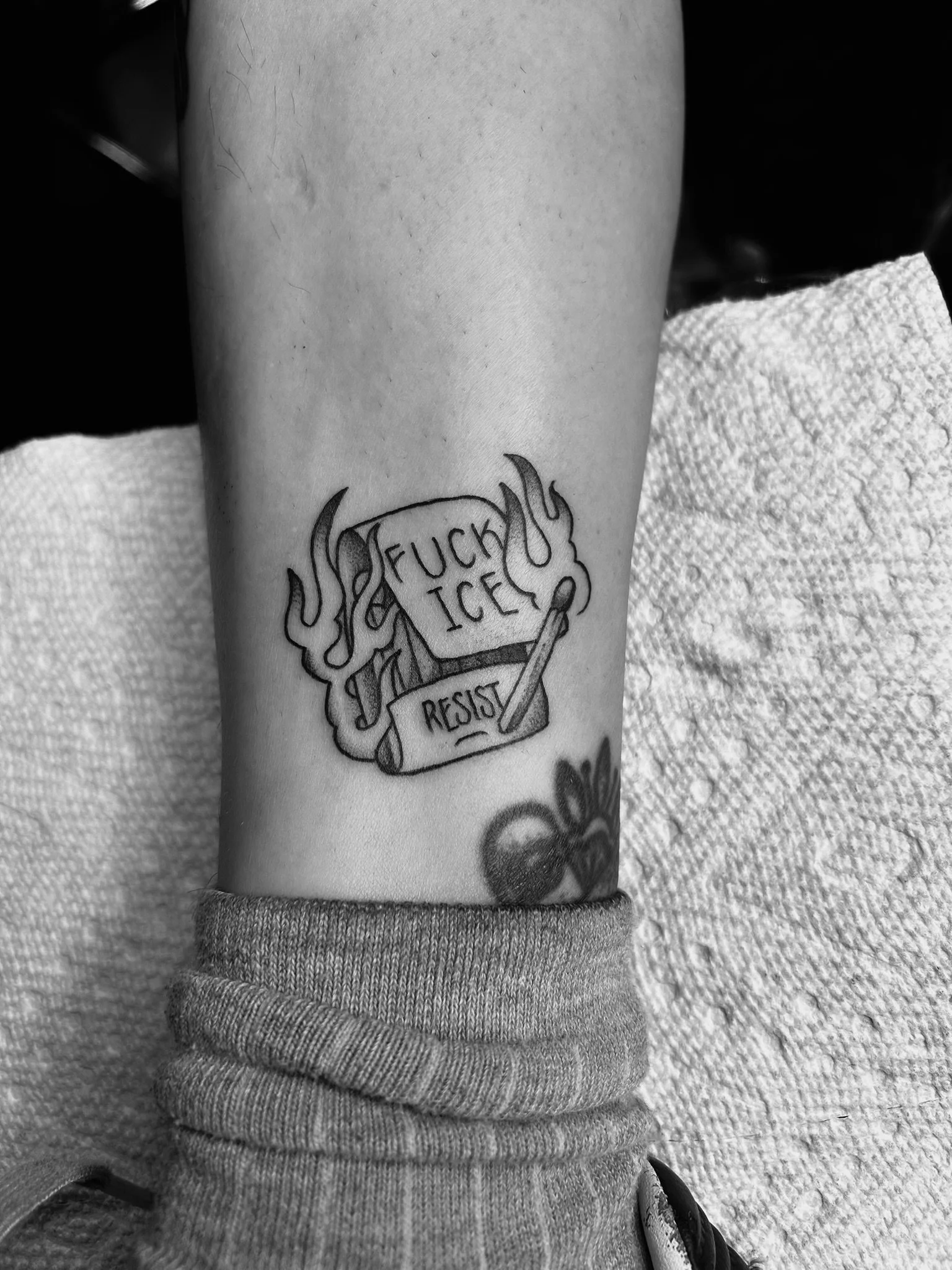 Black-and-white photo of a forearm tattoo featuring flames, the words “FUCK ICE,” and a scroll reading “RESIST.”