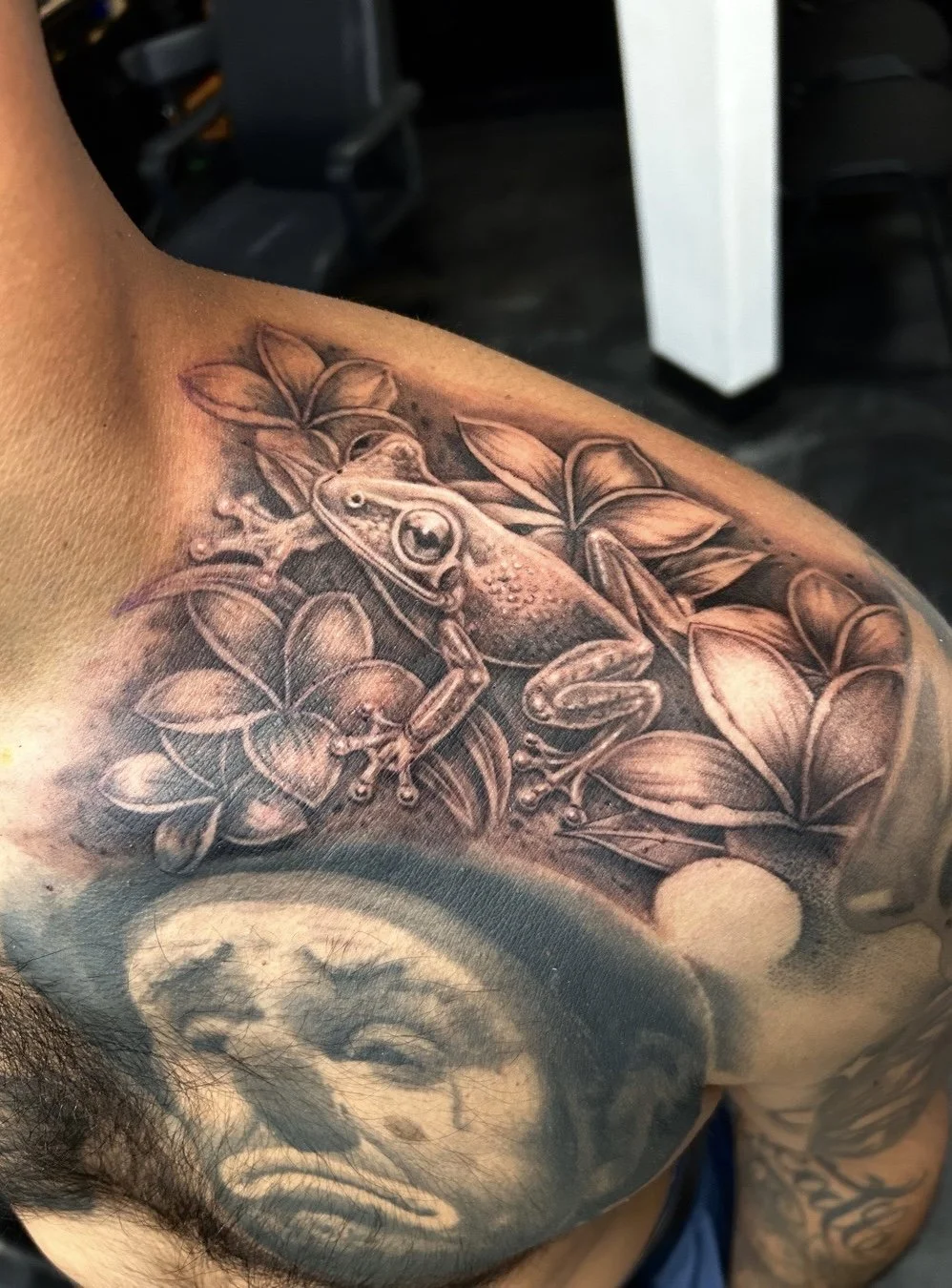Close-up of a detailed frog and floral tattoo on a person’s upper arm, with a portrait tattoo below