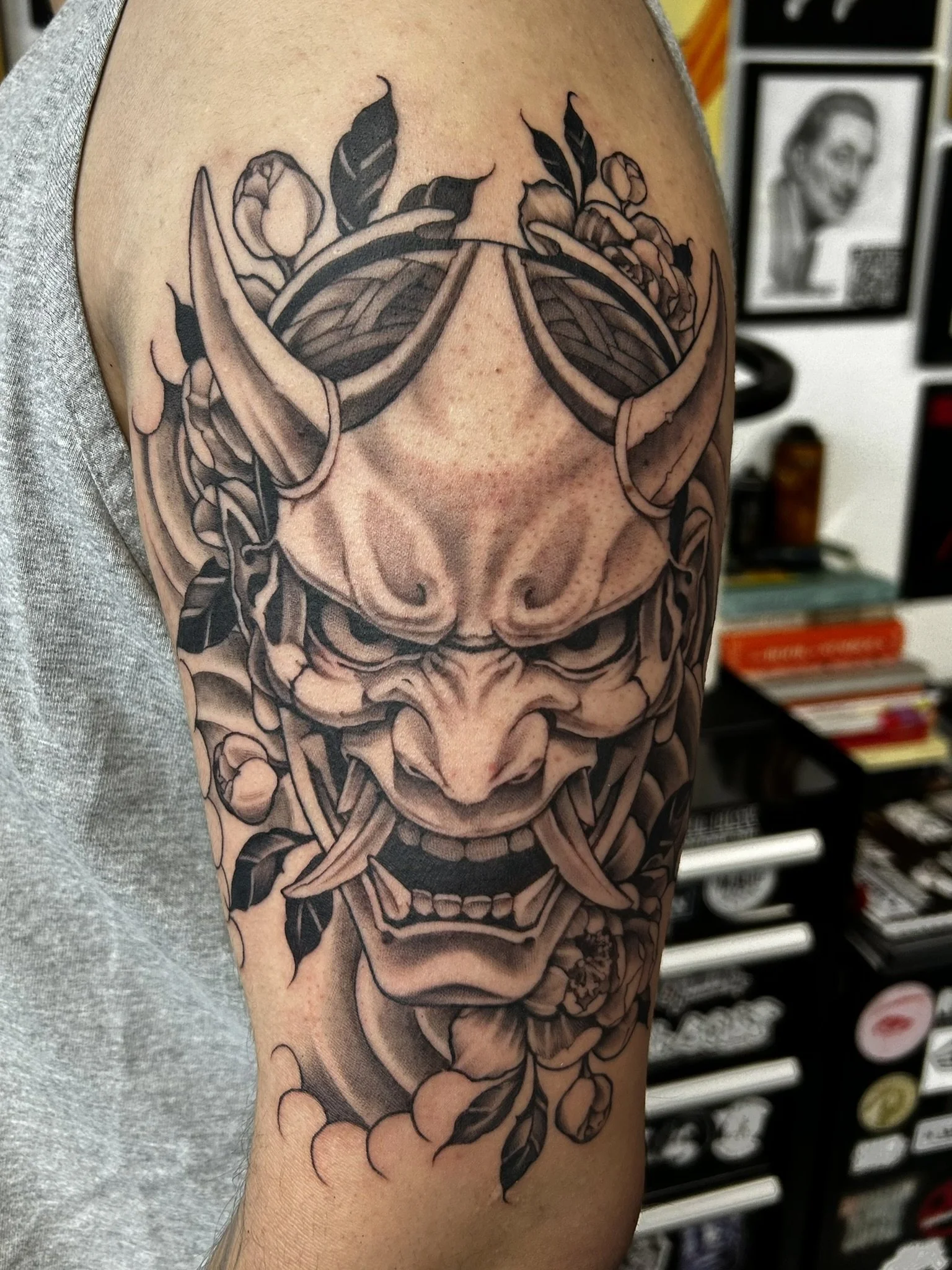 Detailed black-and-gray Japanese oni mask tattoo surrounded by flowers and leaves on an upper arm.