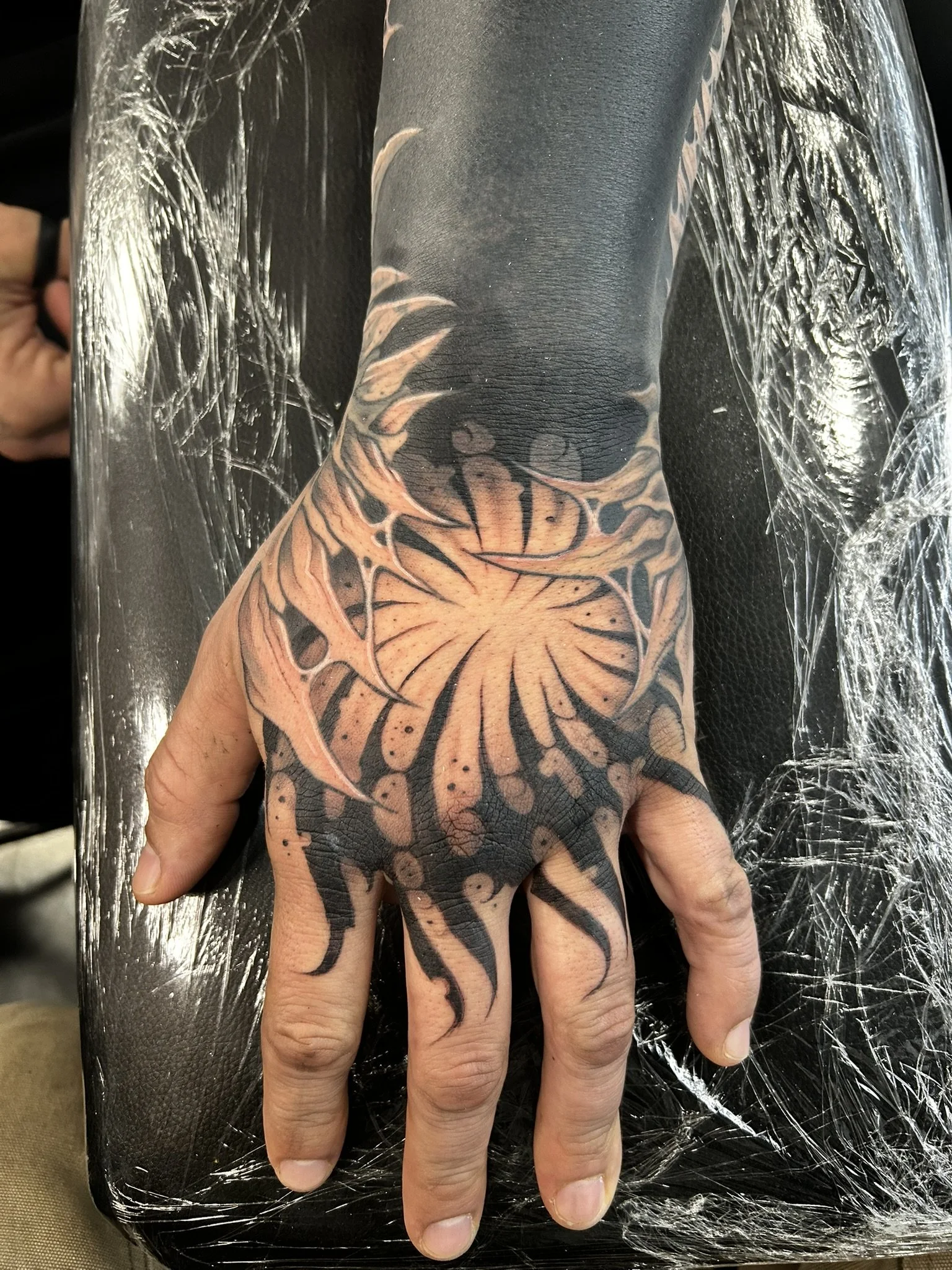Close-up of a heavily tattooed hand and forearm with an orange-and-black floral design resting on a black plastic-covered surface