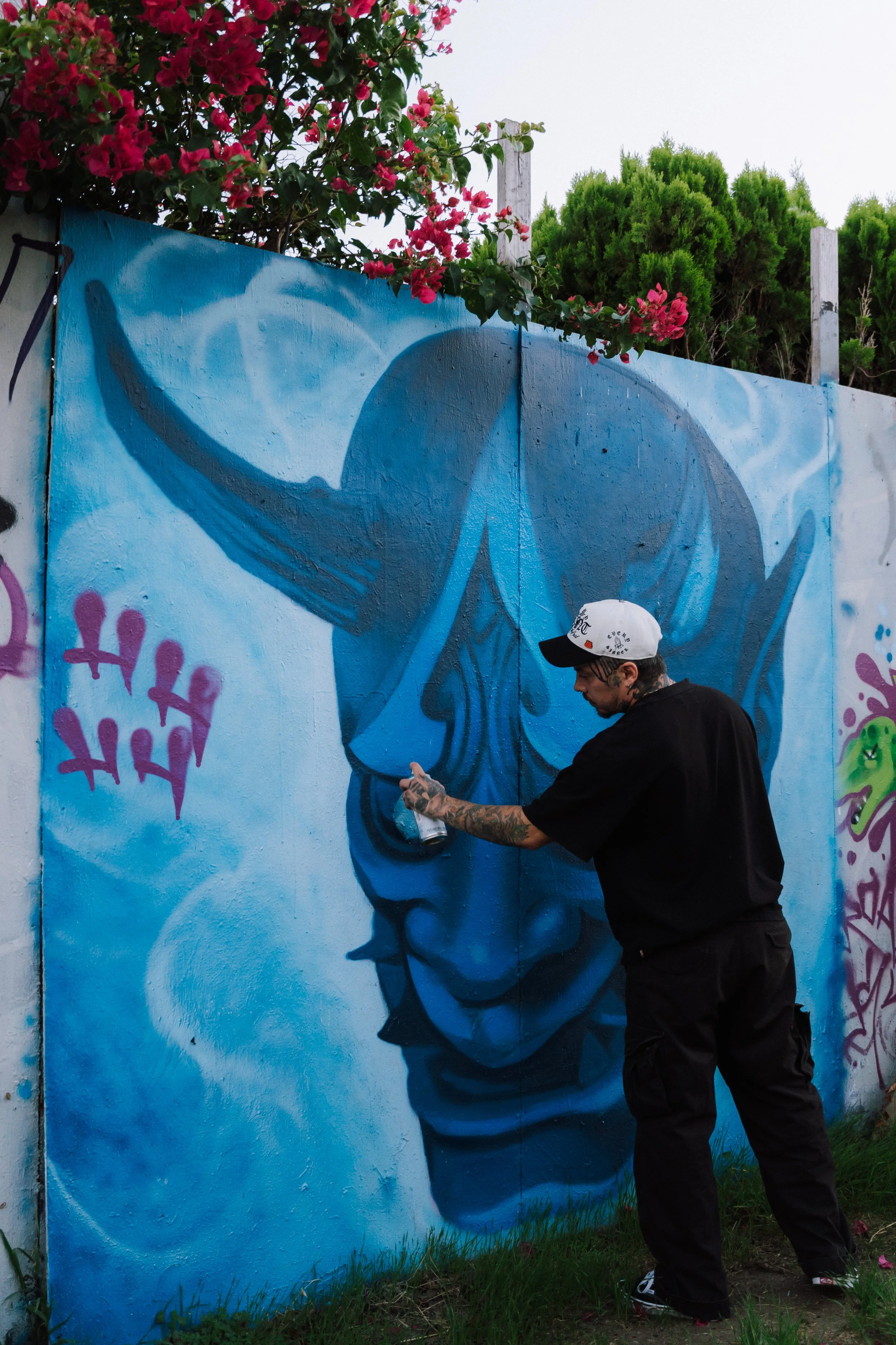 Tattooed graffiti artist spray-painting a blue mural on a wall beneath red bougainvillea flowers.