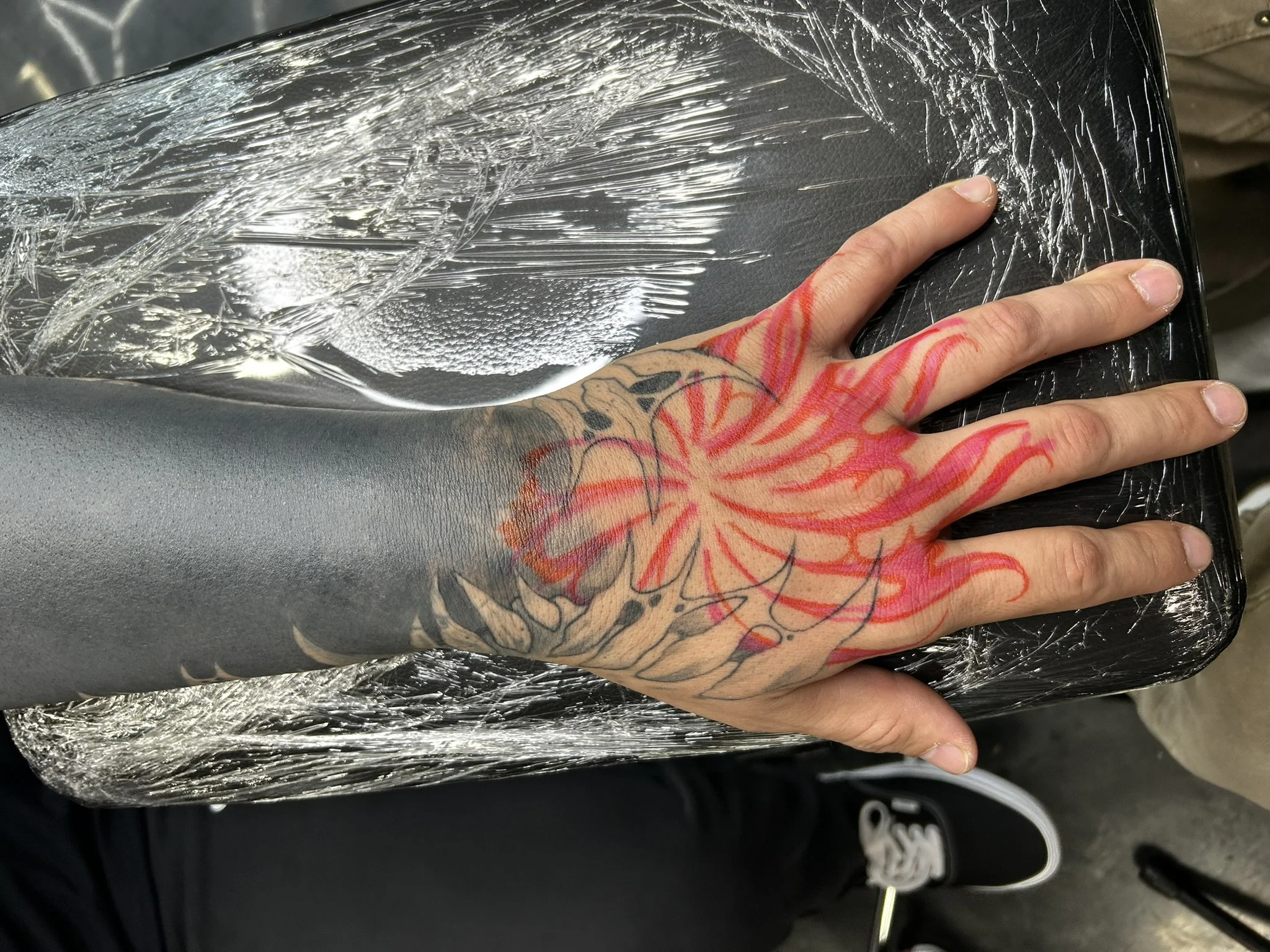 Tattooed hand resting on a plastic-wrapped black leather surface with red stencil lines visible on the skin