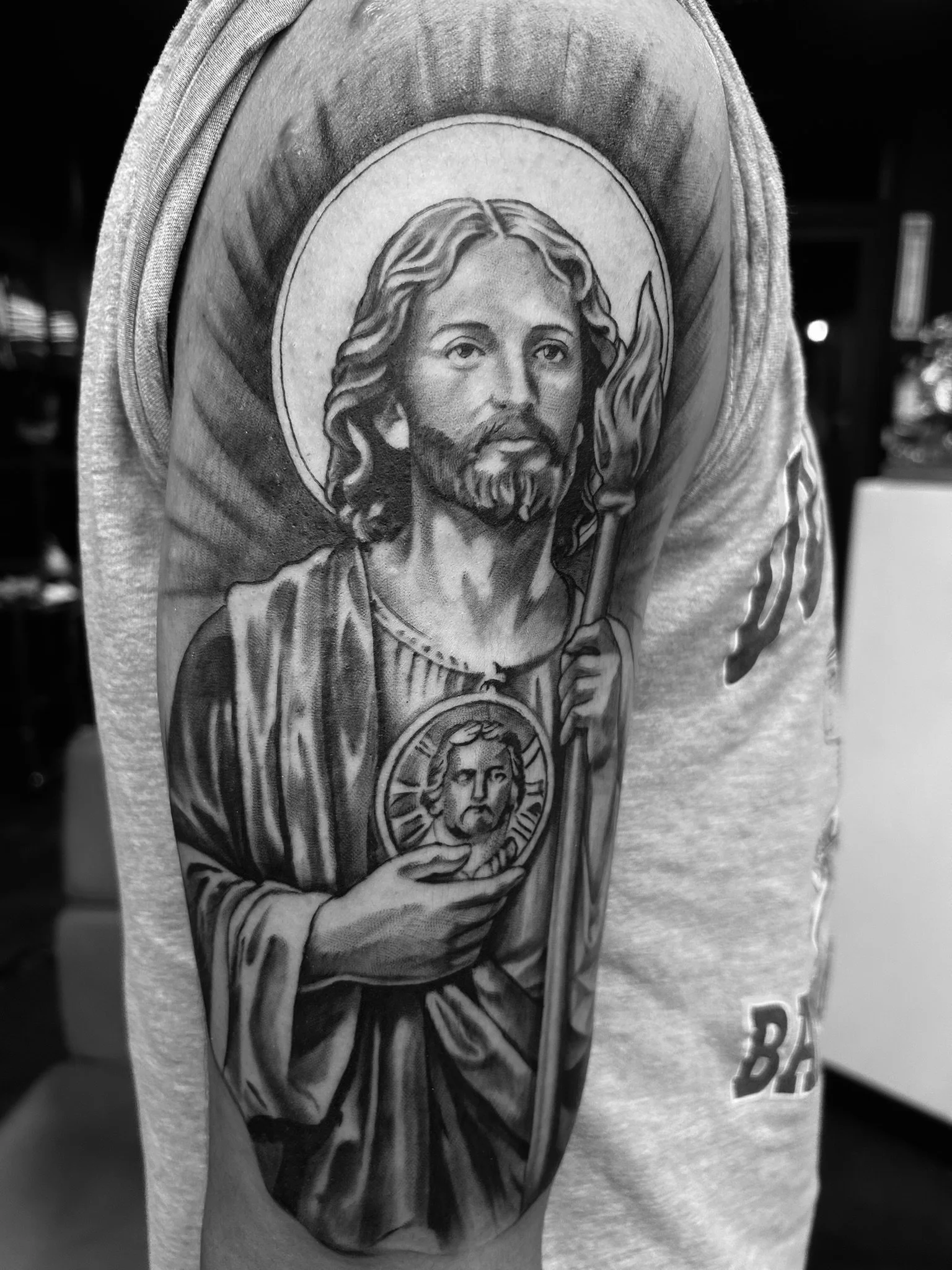 Black-and-white photo of a tattoo depicting Jesus holding a flaming heart and a medallion, surrounded by religious imagery.