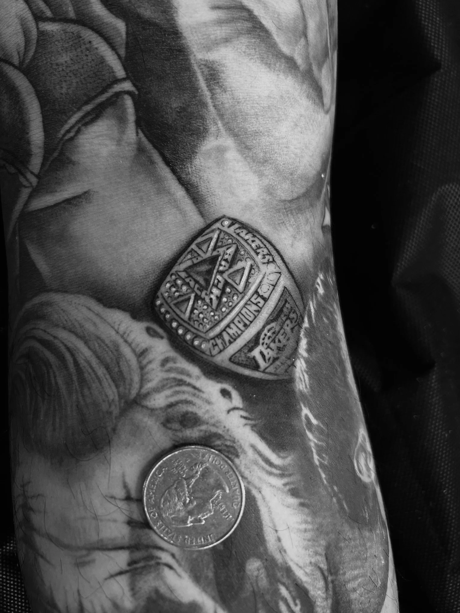 Black-and-white close-up of an arm featuring detailed tattoos, including a heraldic crest, animal, and coin design.