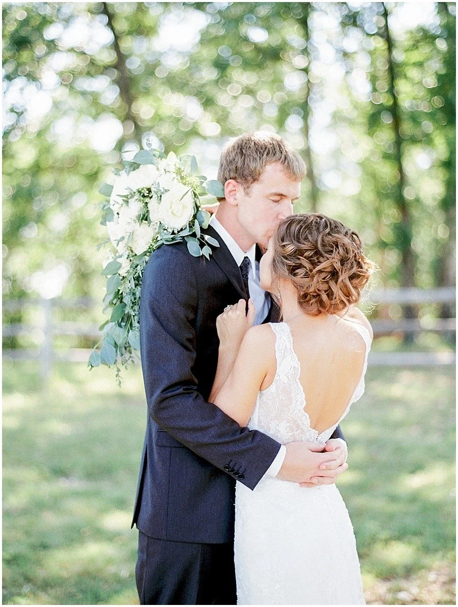 A bride and groom stand close together beneath trees.