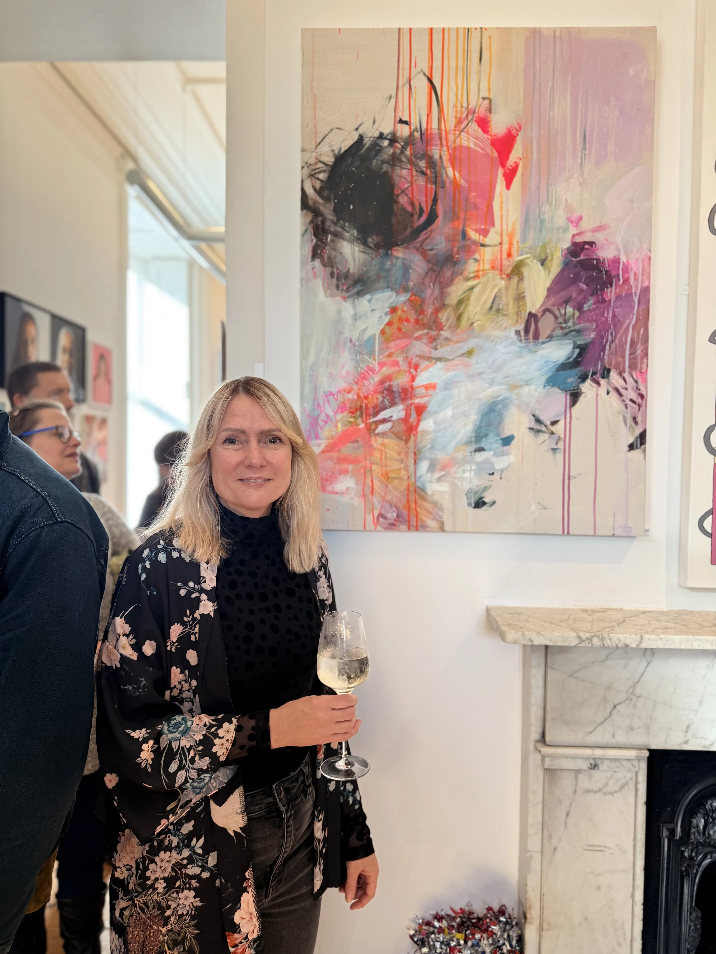 Woman holding a wine glass beside a large colorful abstract painting at an art gallery event