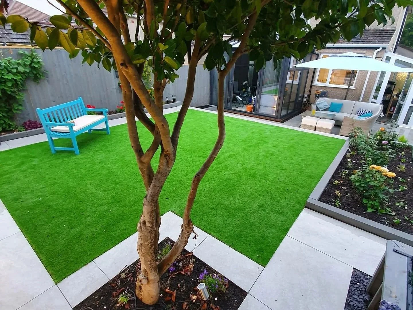 Modern backyard garden with artificial grass, paved patio, tree, flower beds, turquoise bench, and outdoor seating area