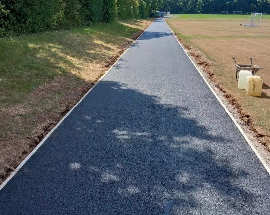 Newly paved asphalt pathway alongside a soccer field, with construction tools and materials nearby