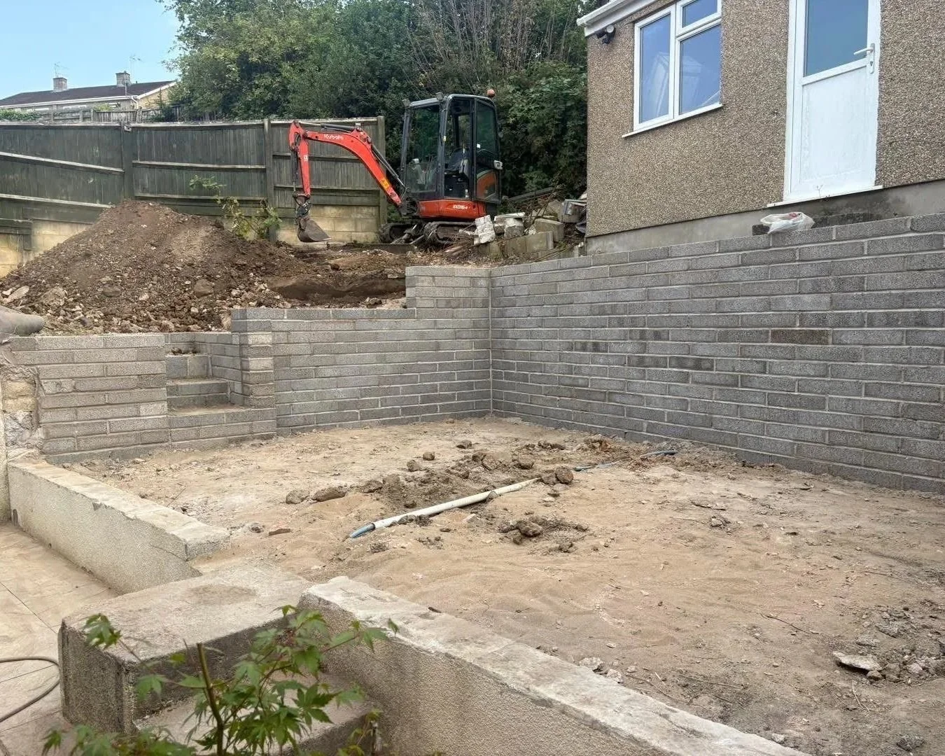 Residential backyard under construction with a compact excavator, exposed soil, and newly built gray block retaining walls.