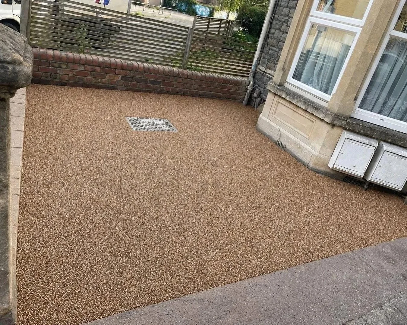 Textured resin-bound driveway beside a stone house with windows, brick wall, drainage grate, and utility covers