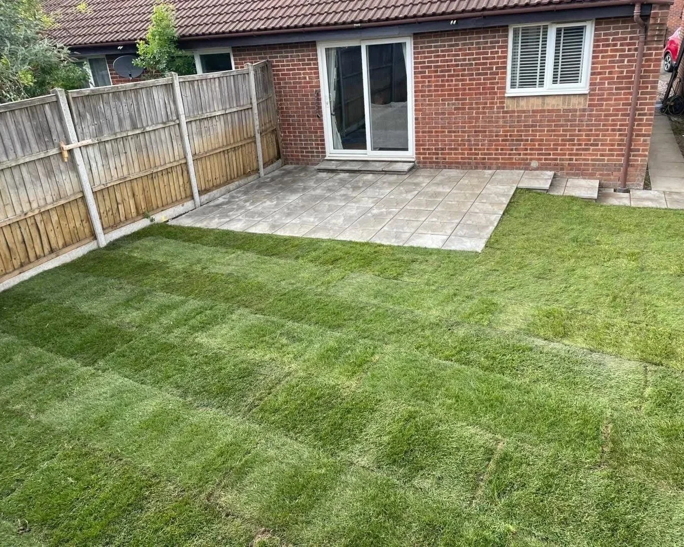 Backyard lawn with a paved patio, wooden fence, and brick house with sliding glass doors