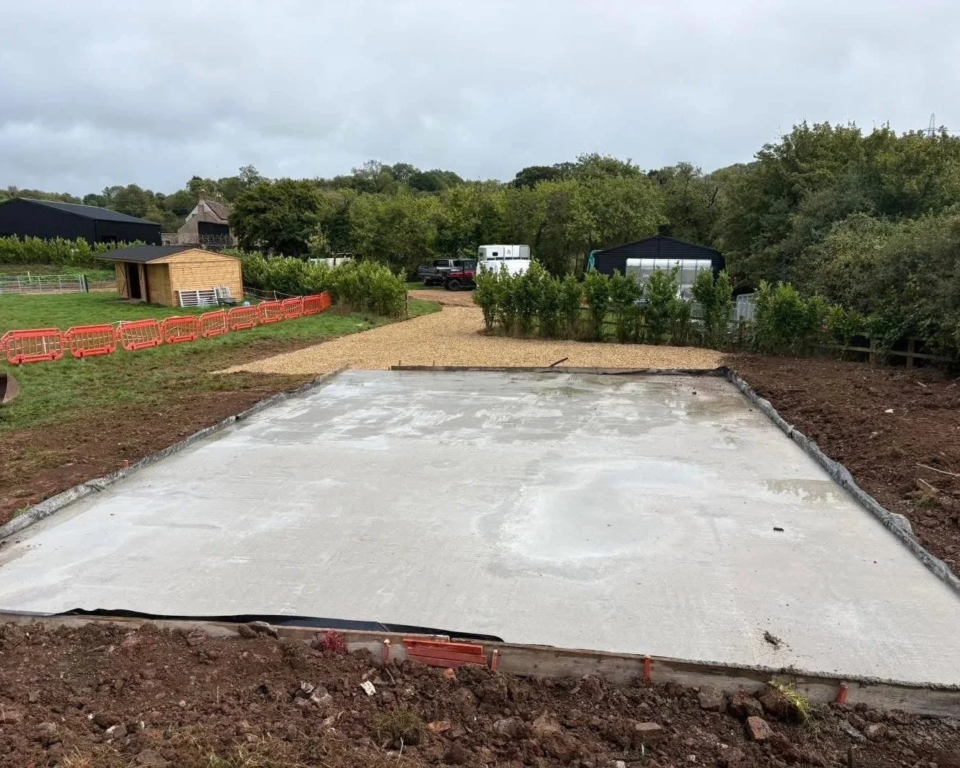 Freshly poured concrete slab under construction in a rural yard, with gravel path, wooden shed, trees, and red safety barriers.