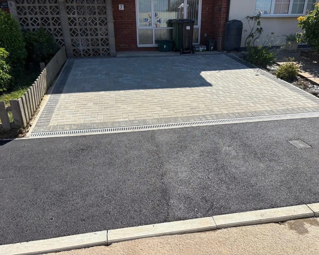 Newly paved residential driveway with interlocking gray pavers, drainage channel, trash bins, and landscaped garden beds
