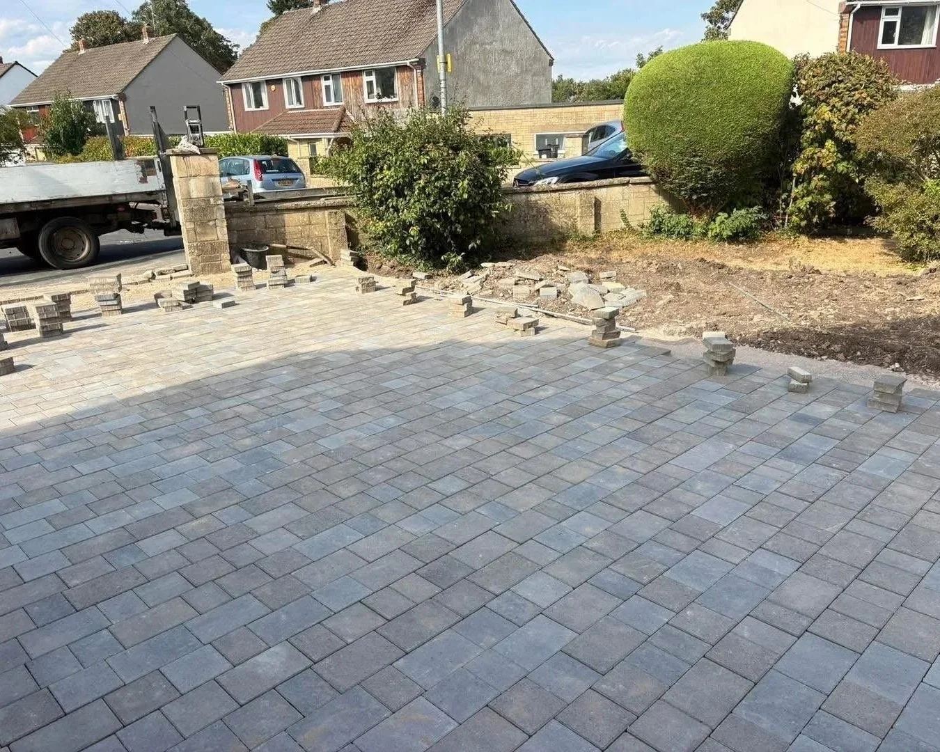 Newly paved residential driveway and patio with gray block pavers, unfinished garden landscaping, hedges, parked cars, and suburban homes