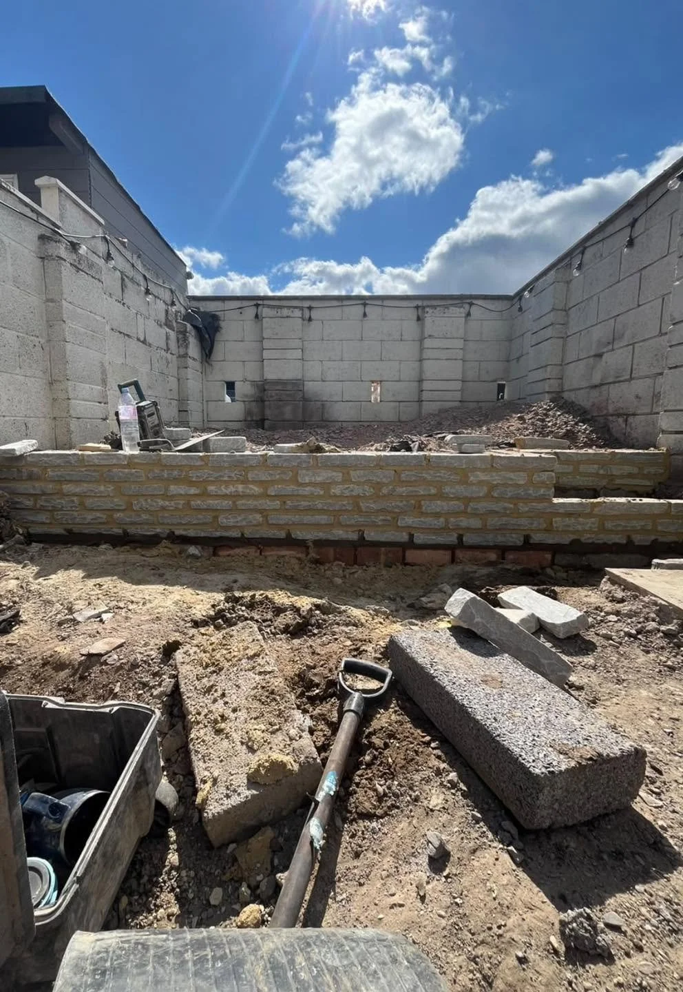 Backyard construction site with a newly built brick retaining wall, cinder block walls, dirt, concrete blocks, and a shovel under a blue sky