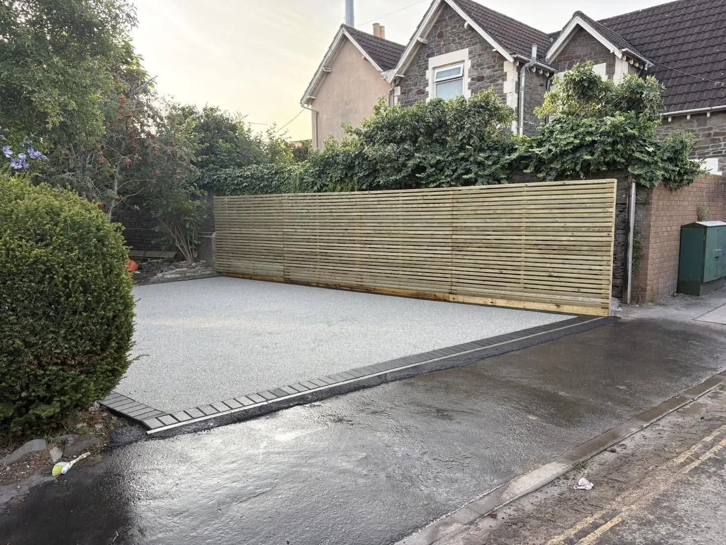 Newly paved residential driveway with a horizontal wood privacy fence, landscaped garden, and stone house after rain