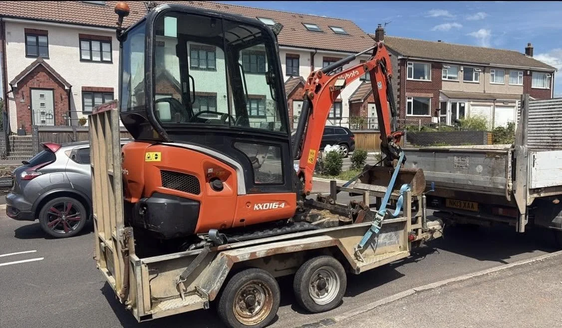 Kubota KX016-4 mini excavator secured on a trailer beside a flatbed truck on a residential street