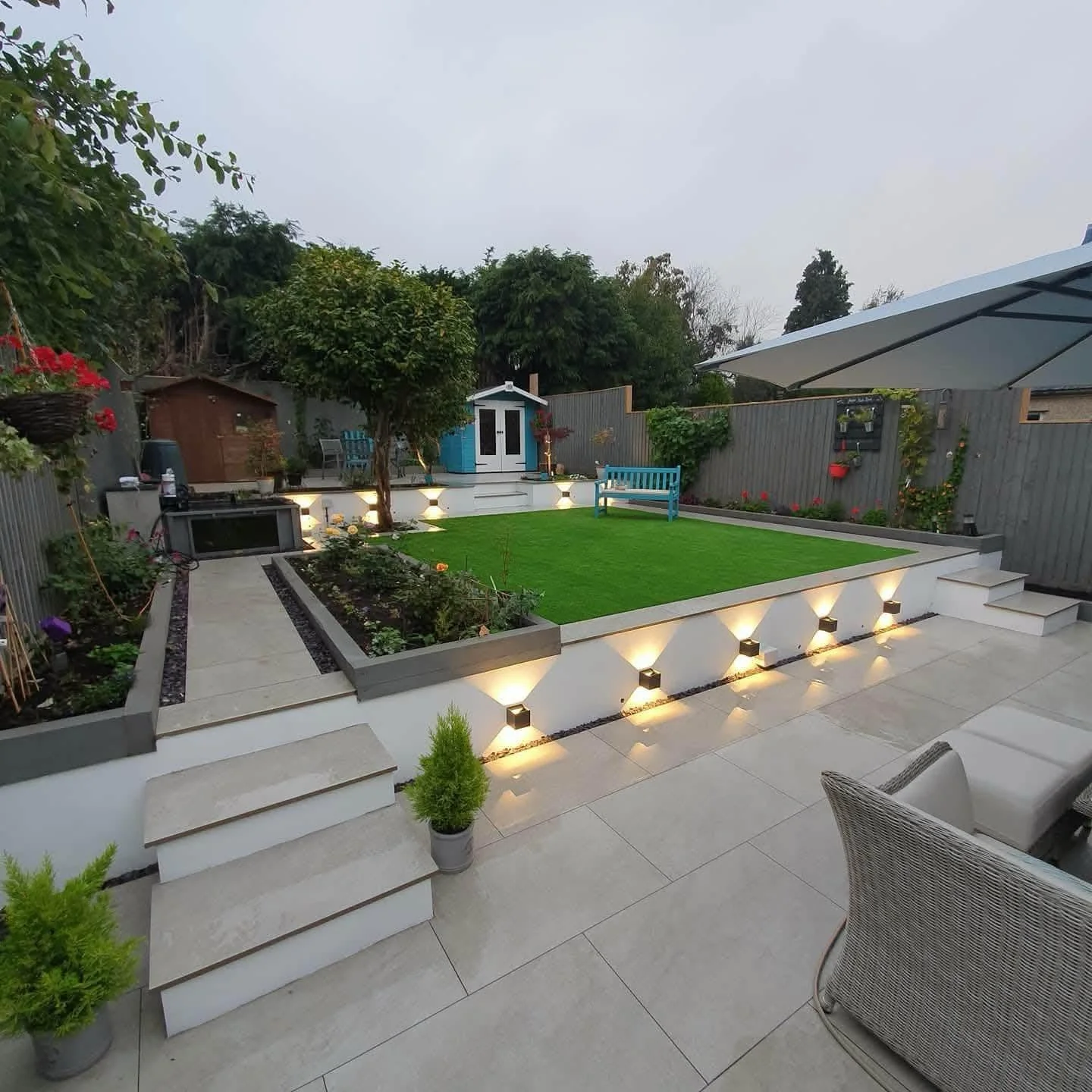 Modern landscaped backyard patio with artificial lawn, raised flower beds, illuminated steps, turquoise bench, garden shed, and glass-roofed seating area