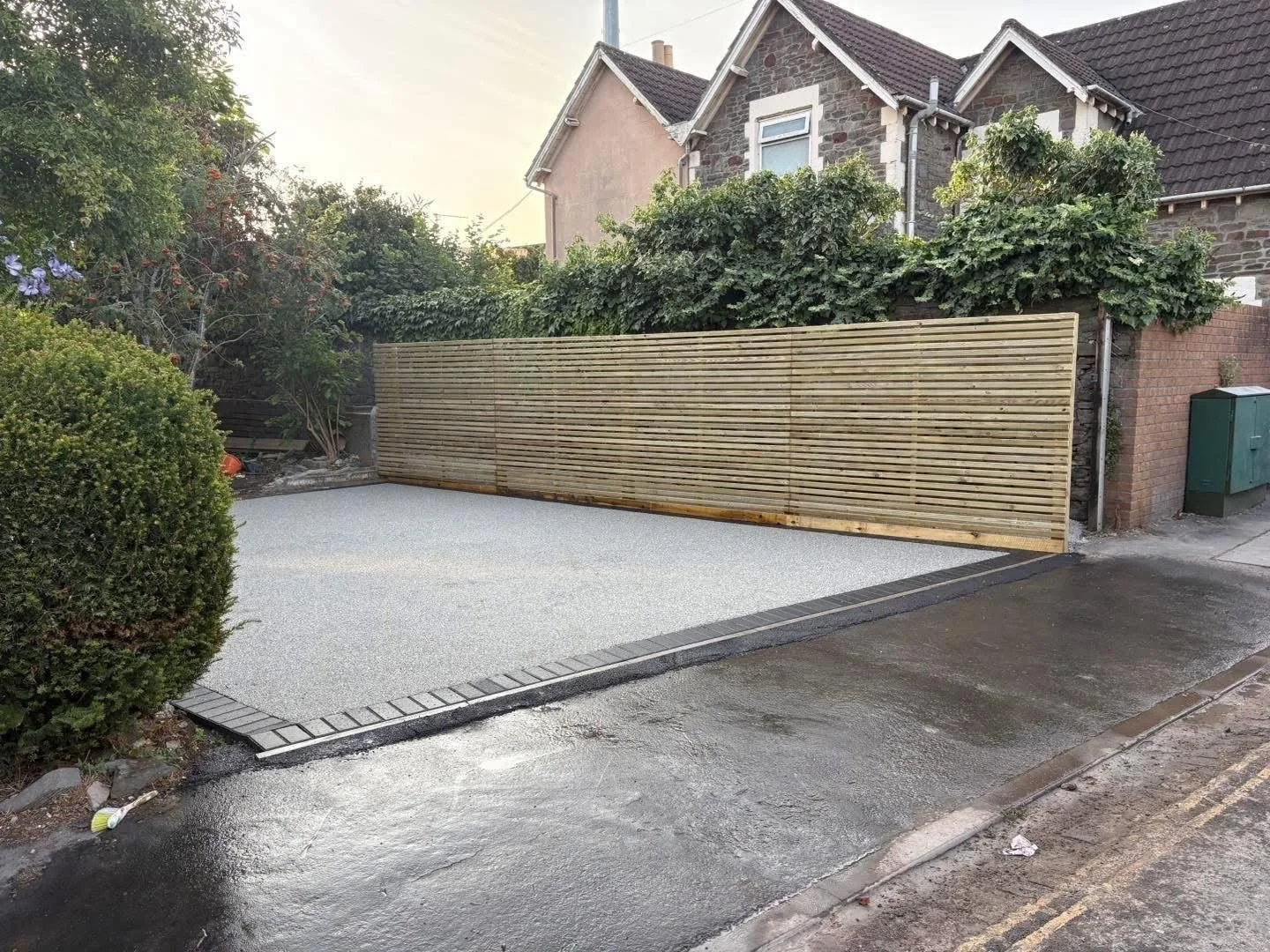 Newly paved residential driveway with a wooden privacy fence, landscaped garden, and stone house