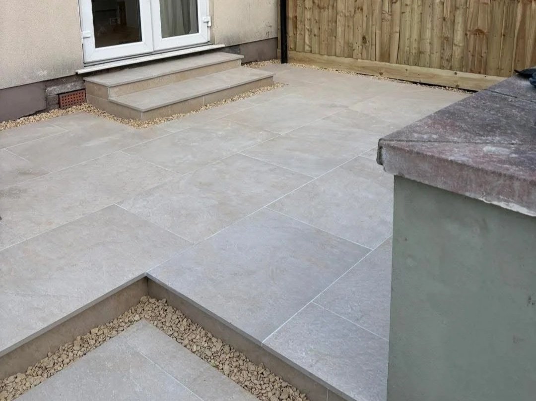 Gray tiled backyard patio with raised steps leading to white French doors, gravel borders, and a wooden privacy fence.