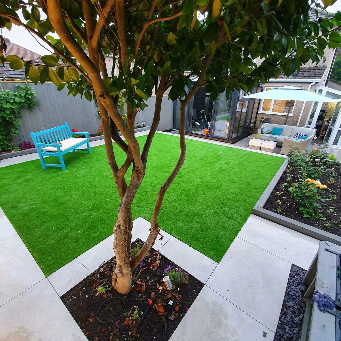 Landscaped backyard with artificial grass, tiled patio, mature multi-trunk tree, blue bench, flower beds, and covered outdoor seating area