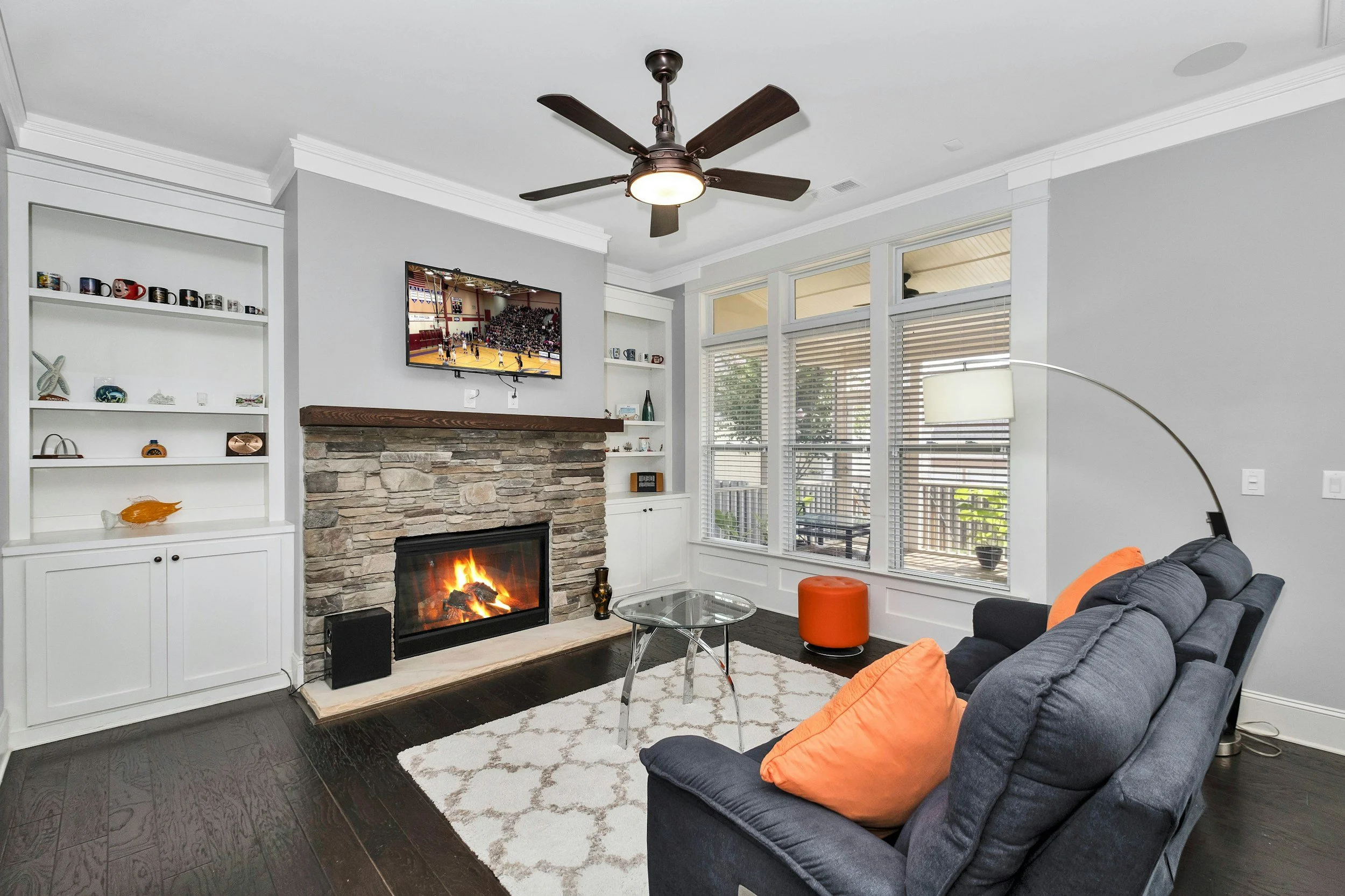 Bright living room with a stone fireplace, mounted TV, ceiling fan, built-in shelves, large windows, glass coffee table, and navy sofa with orange pillows.