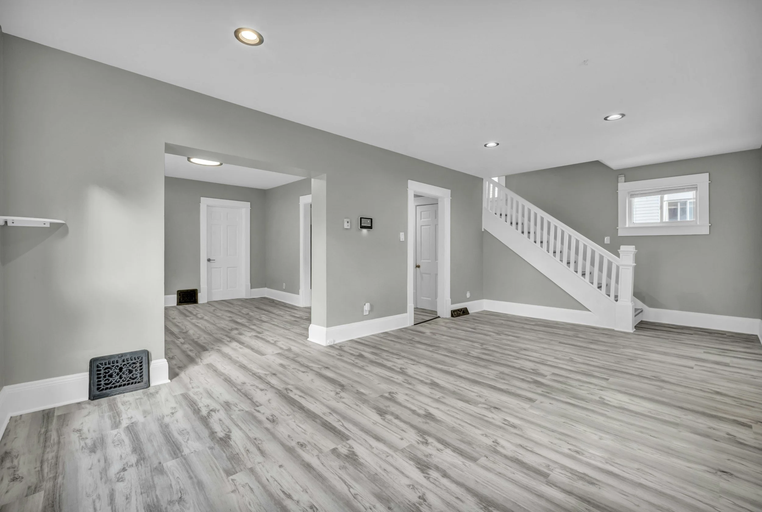 Spacious renovated living room with gray walls, white trim, gray wood-look flooring, recessed lighting, staircase, doors, and window