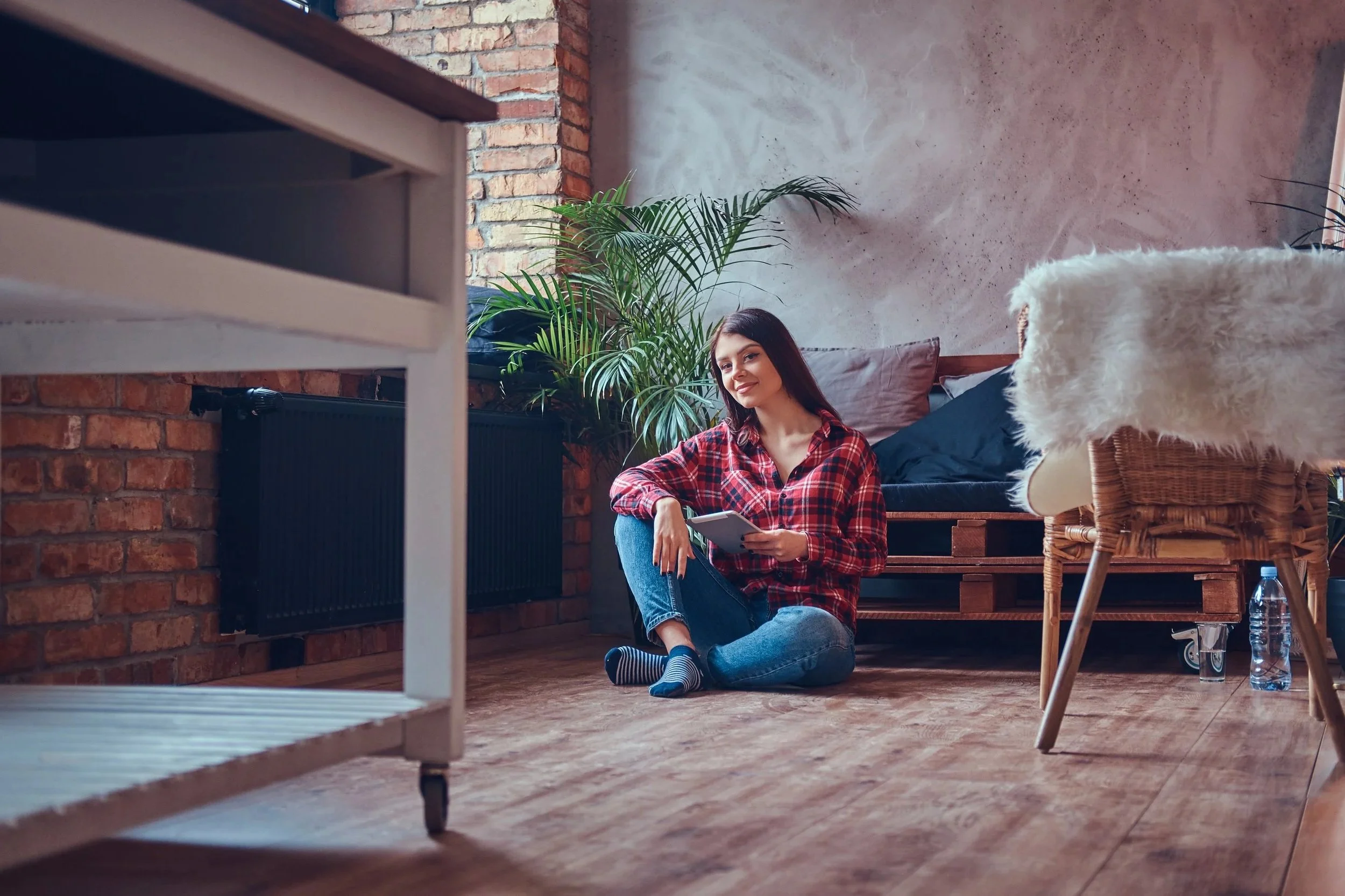 Woman in a red plaid shirt sitting on a wooden floor and reading in a cozy brick-walled room