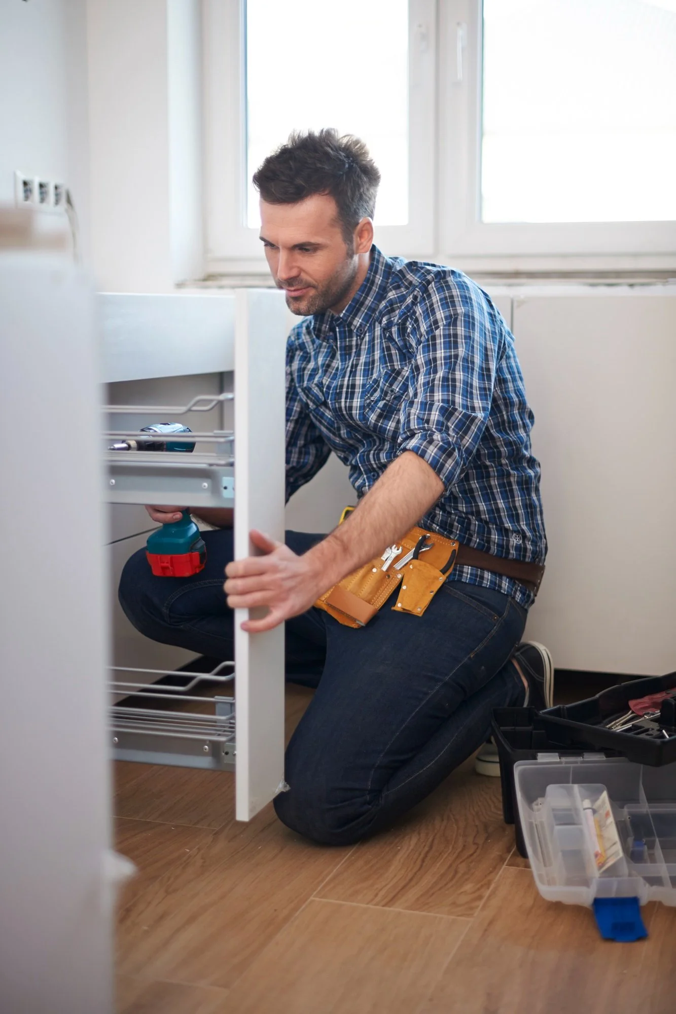 Man installing a pull-out kitchen cabinet drawer while kneeling with tools nearby