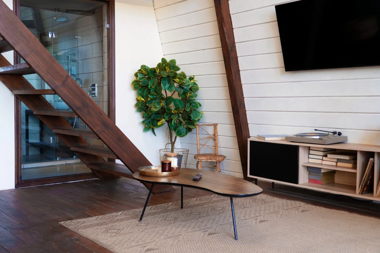 Modern living room with a wooden staircase, indoor tree, coffee table, turntable, books, and wall-mounted television