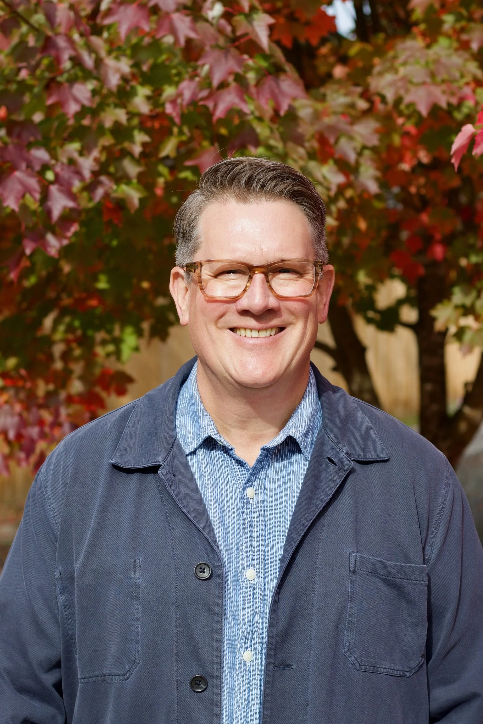 Troy S. Anderson, church and nonprofit lawyer, smiling and wearing glasses and a navy jacket standing in front of autumn foliage