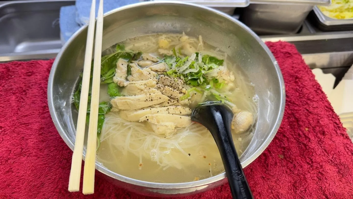 Vietnamese-style chicken rice noodle soup with herbs, mushrooms, chopsticks, and a black spoon in a metal bowl