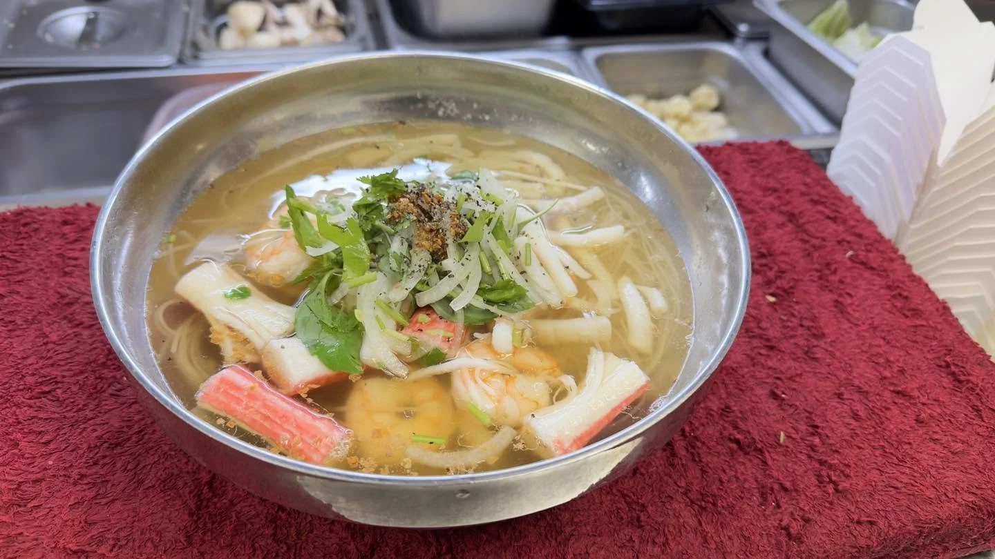 Metal bowl of seafood noodle soup with crab sticks, shrimp, mushrooms, herbs, and sliced vegetables