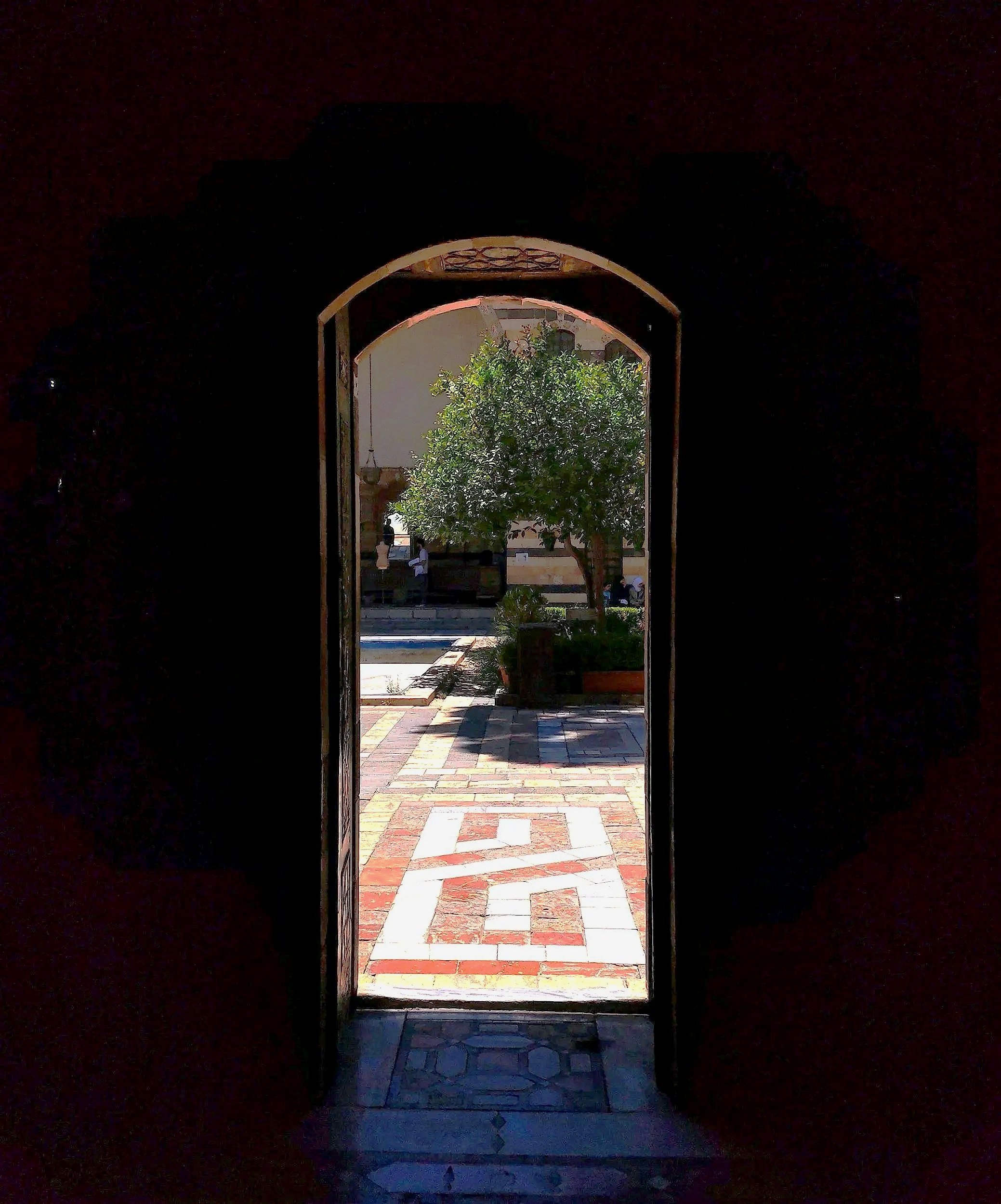 Sunlit courtyard viewed through an arched doorway, with a potted citrus tree, tiled walkway, and small fountain.