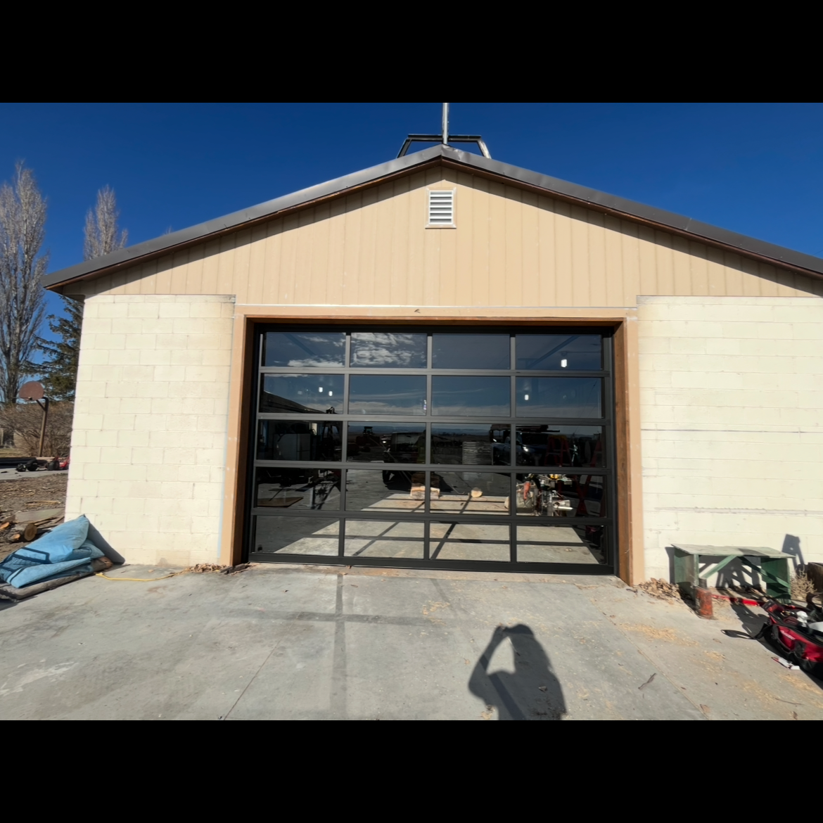 Front view of a tan workshop or garage with a large black-framed glass garage door under a clear blue sky.