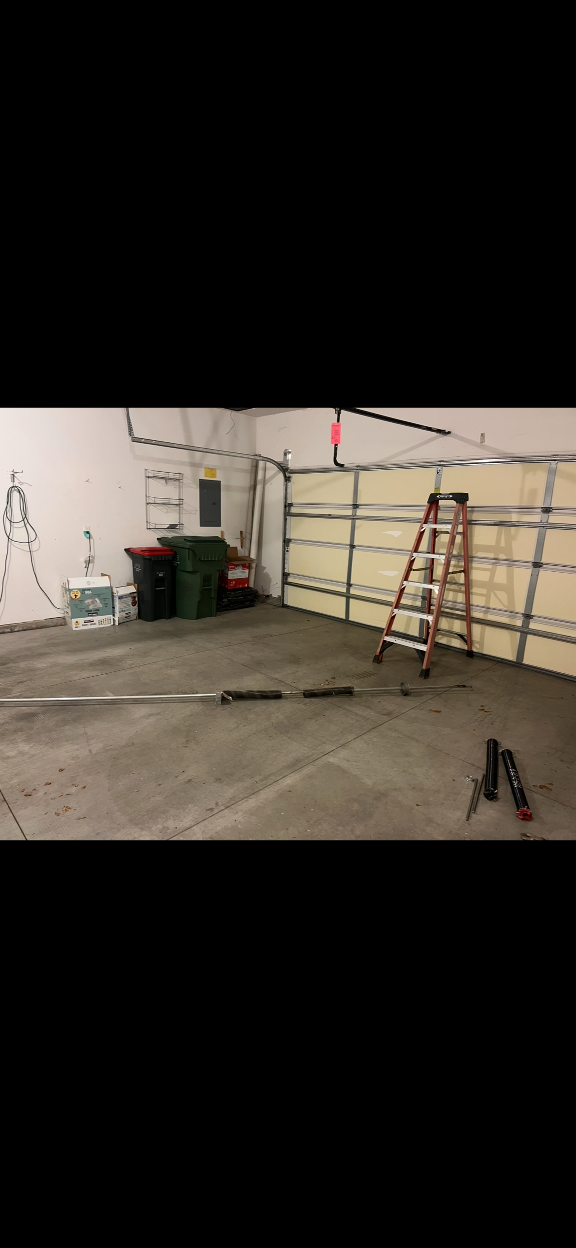 Garage interior with a closed sectional door, red step ladder, trash bins, storage shelves, and detached garage door hardware on the concrete floor.