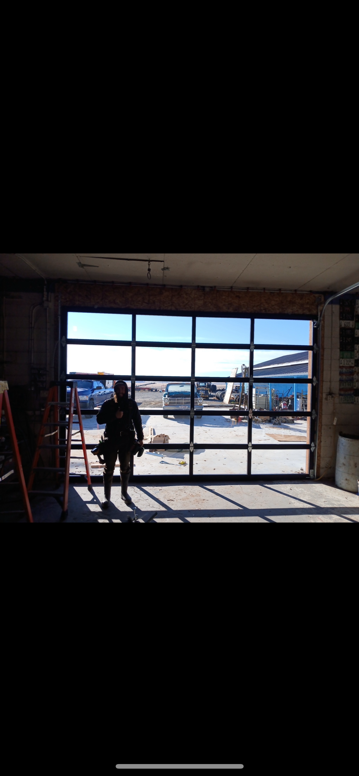 Worker standing inside a garage with a ladder, looking through a glass-paneled garage door at a vintage pickup truck and industrial yard
