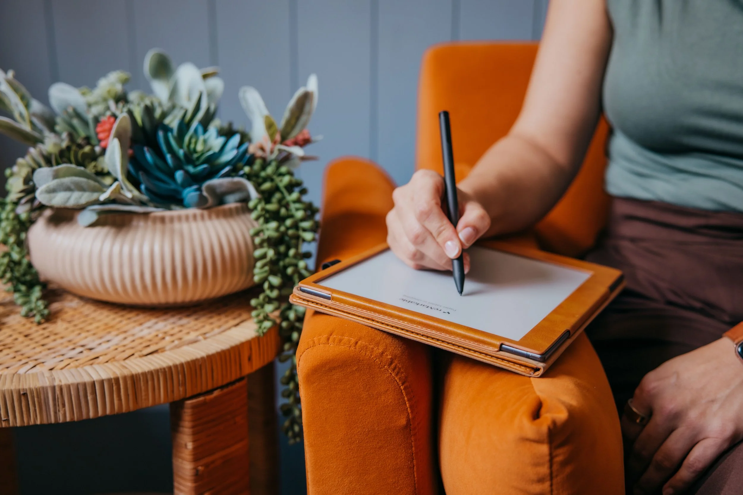Person writing on a tablet while sitting in an orange chair beside a potted succulent arrangement