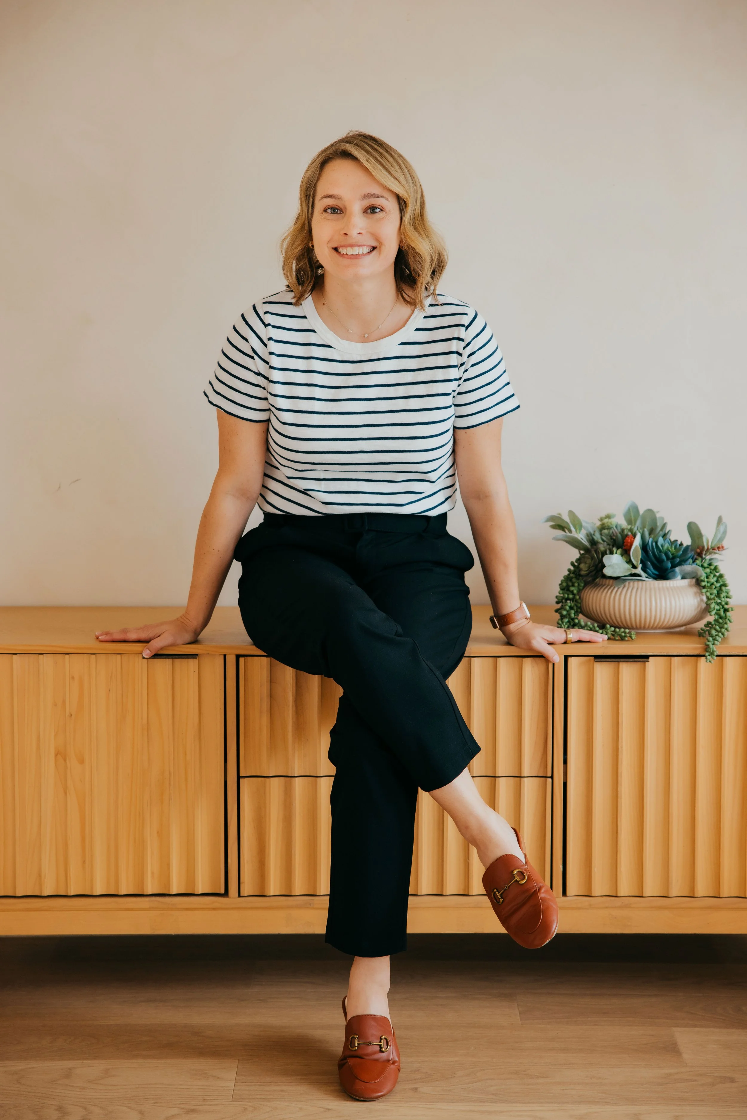 Smiling woman in a striped shirt and black pants sitting on a wooden cabinet beside a potted succulent plant.