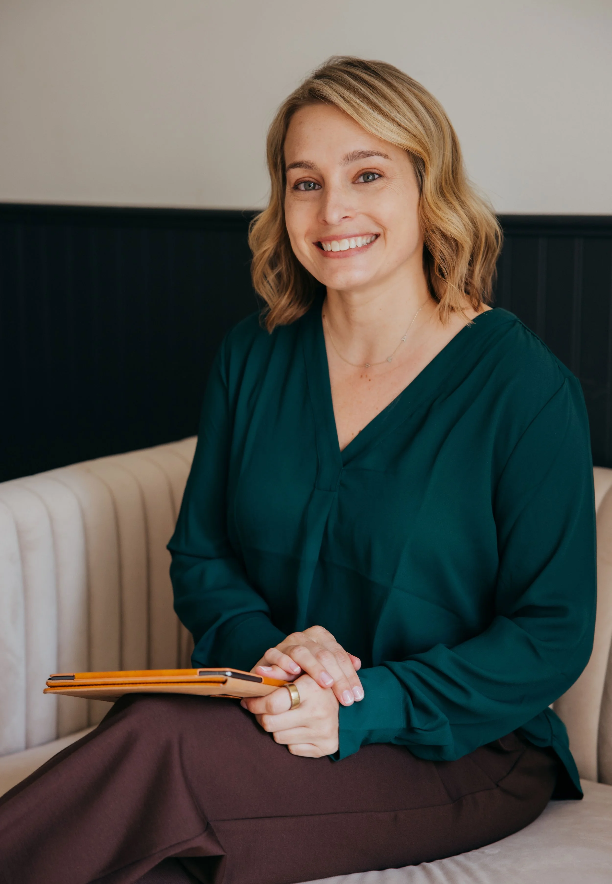 Smiling woman with blonde hair holding a notebook and pen while sitting on a couch