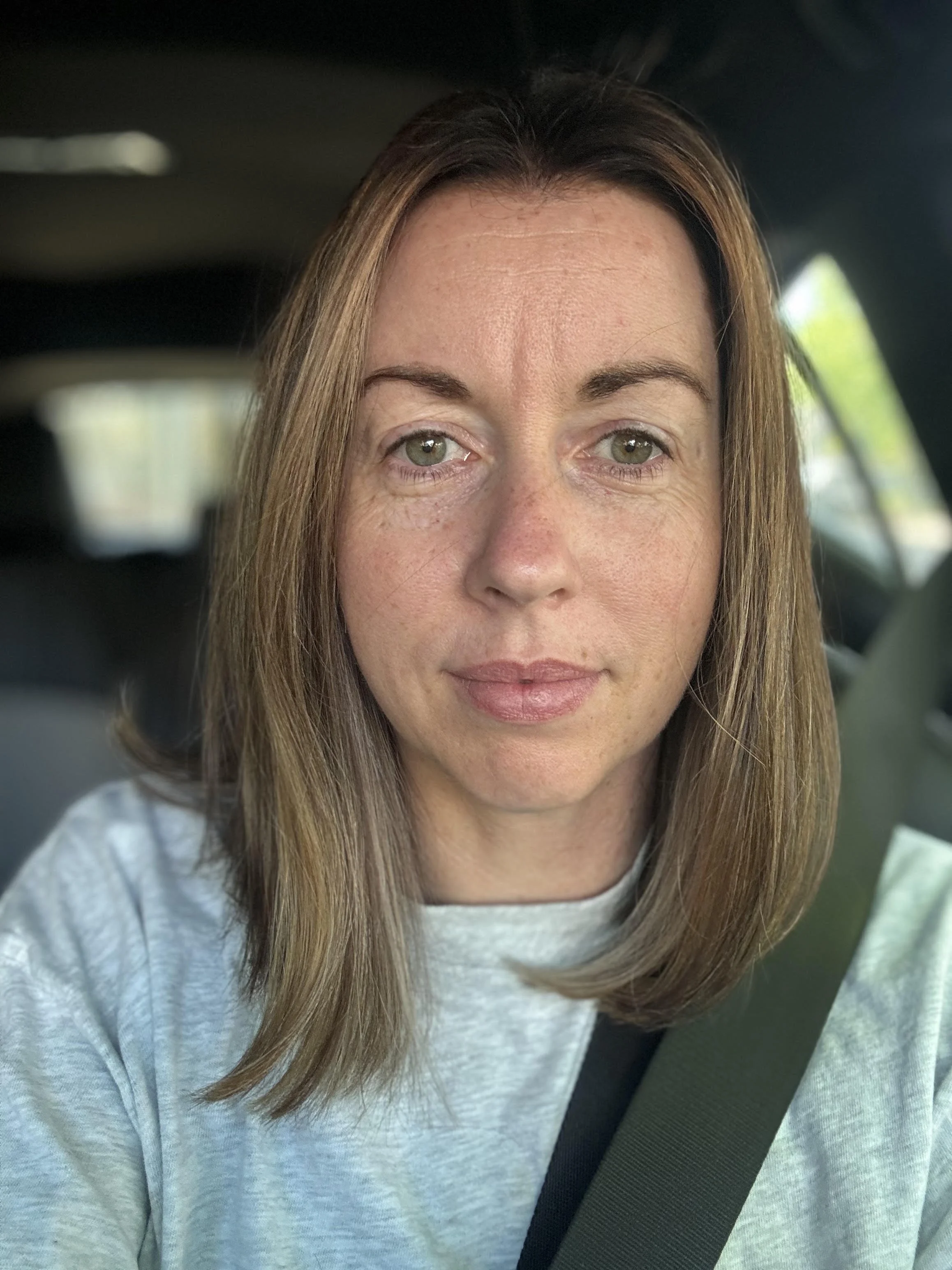 Close-up portrait of a woman with long brown hair and green eyes wearing a seat belt in a car