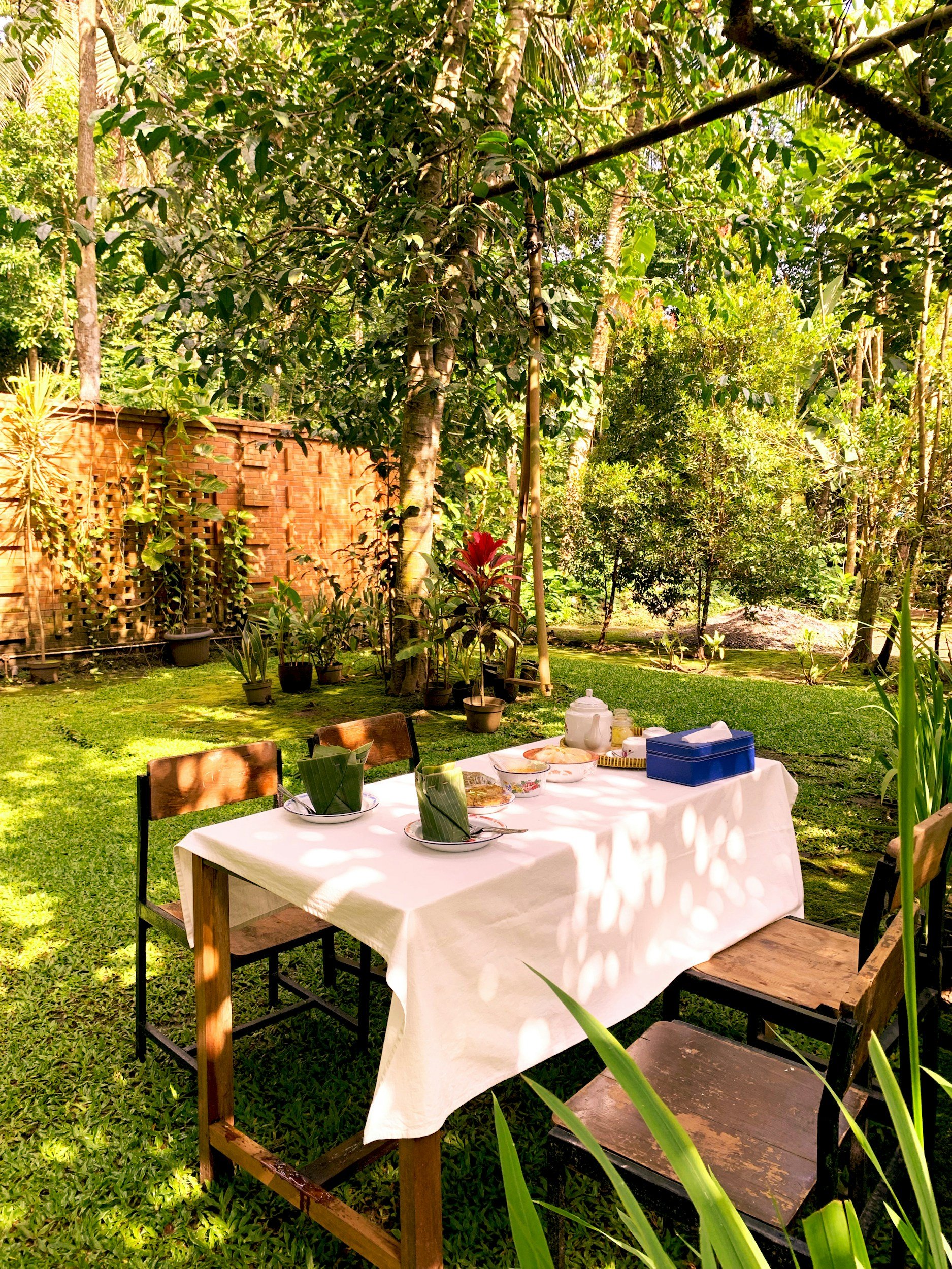 Outdoor dining table set for a meal in a lush tropical garden with potted plants, trees, and a brick wall.