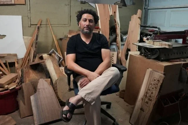 Hamza Arman sitting in a workshop chair surrounded by woodworking tools and lumber