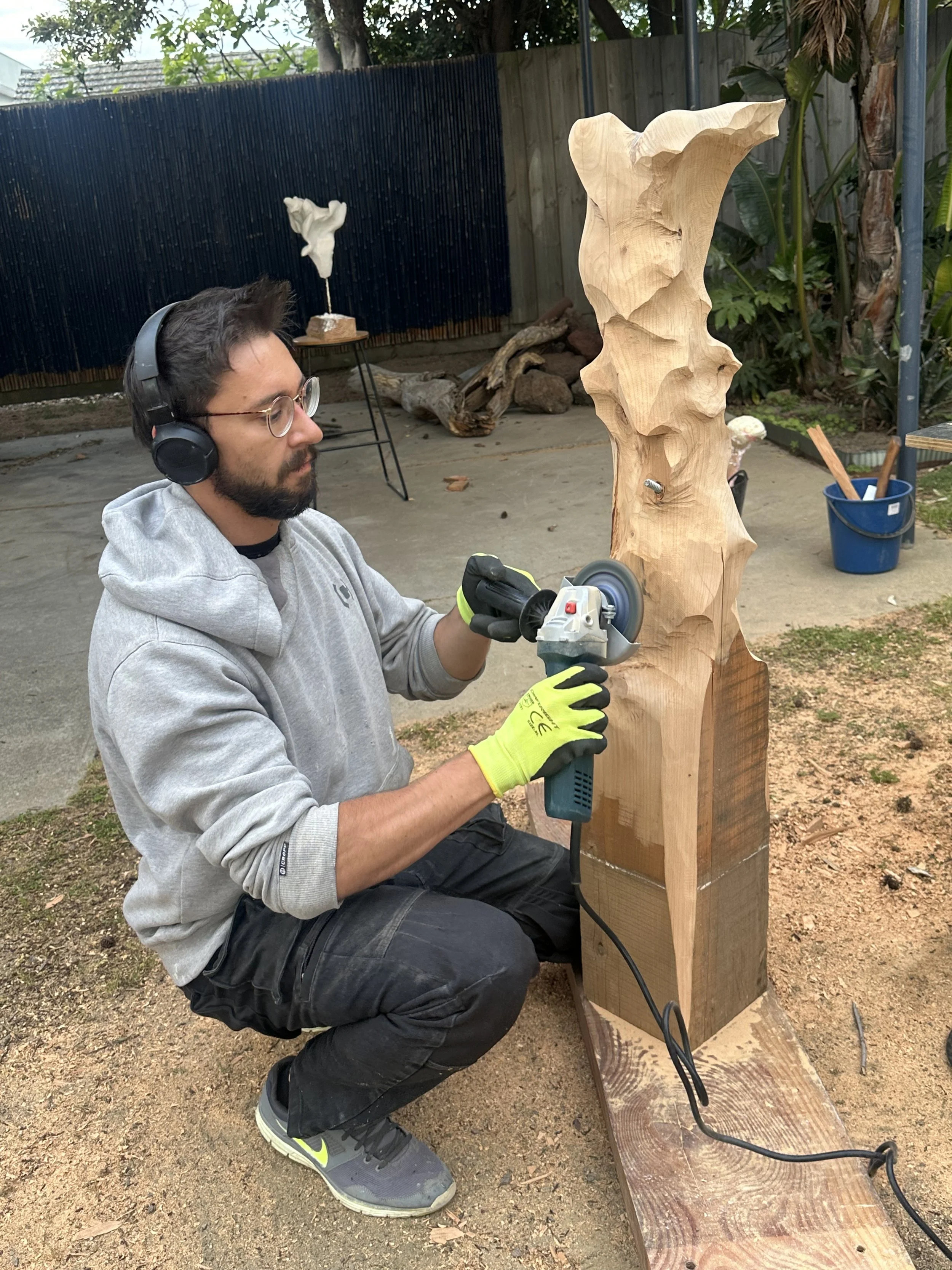 Woodworker wearing safety glasses, headphones, and gloves uses an angle grinder to carve a large wooden sculpture outdoors.