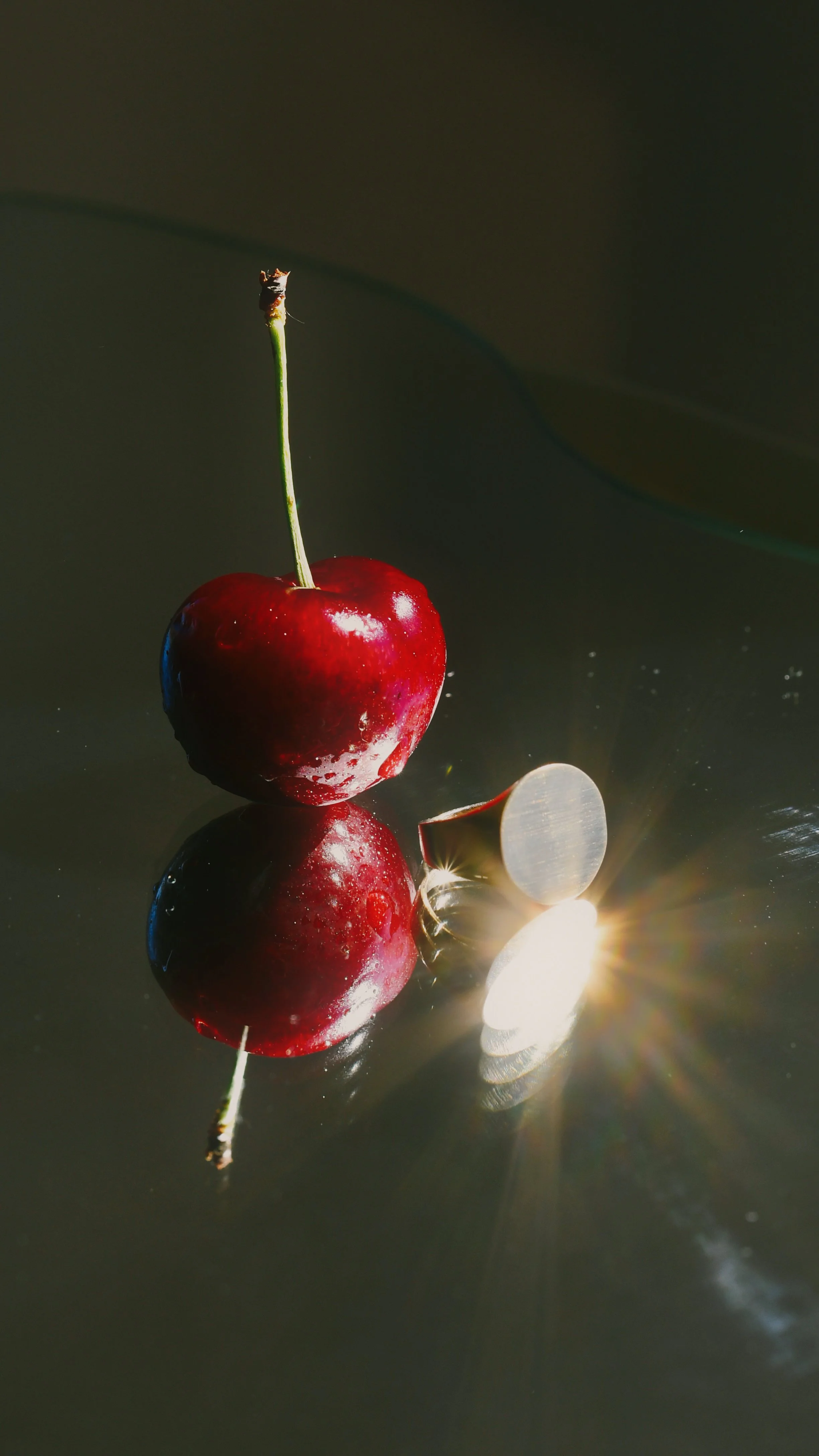 Close-up of two glossy red cherries with stems beside a silver ring on a reflective dark surface